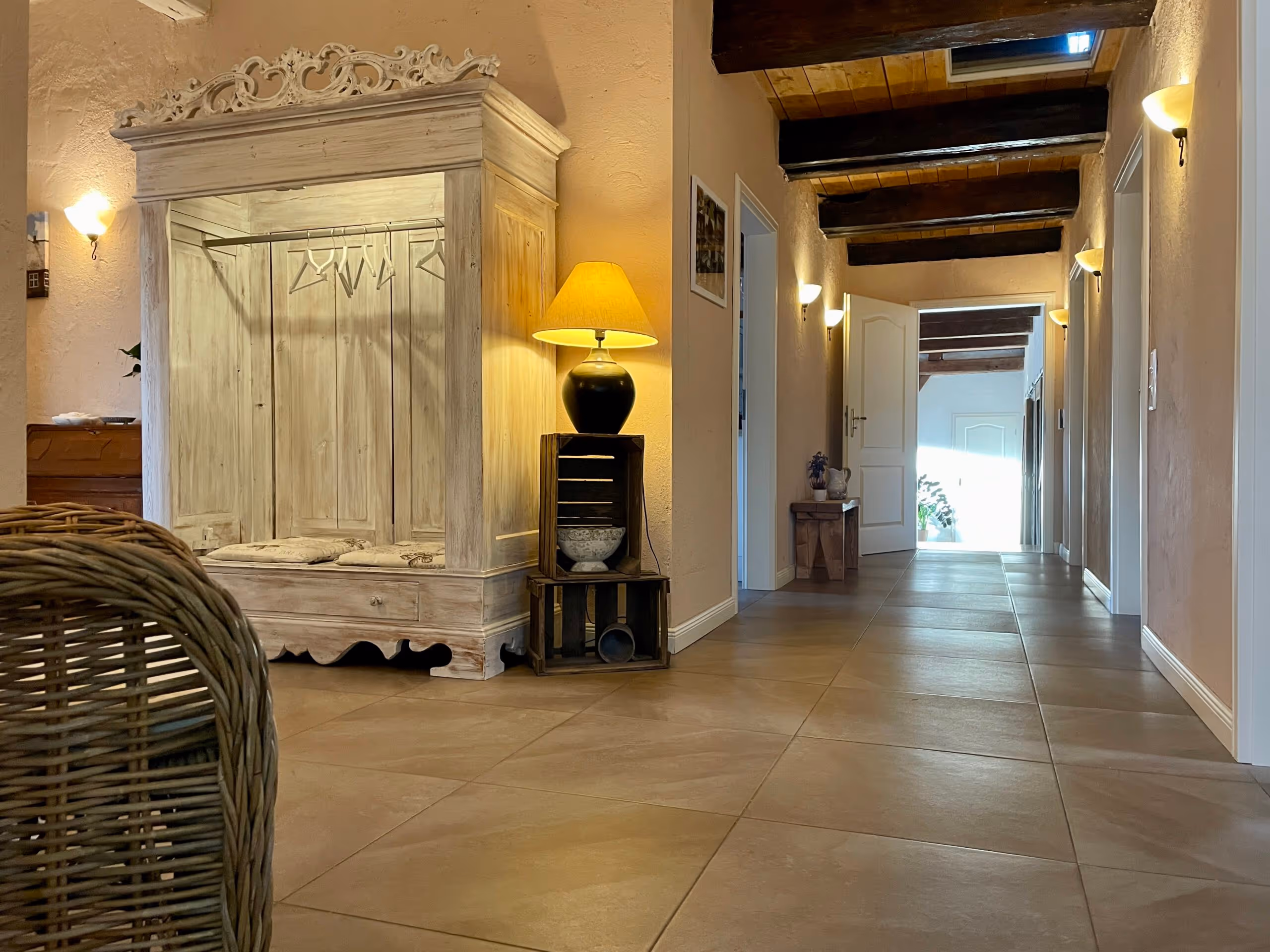 Flur mit cremefarbenen Wänden, Holzbalkendecke und großem weißen Holzschrank mit Sitzkissen links, beleuchtet von Tisch- und Wandlampen.