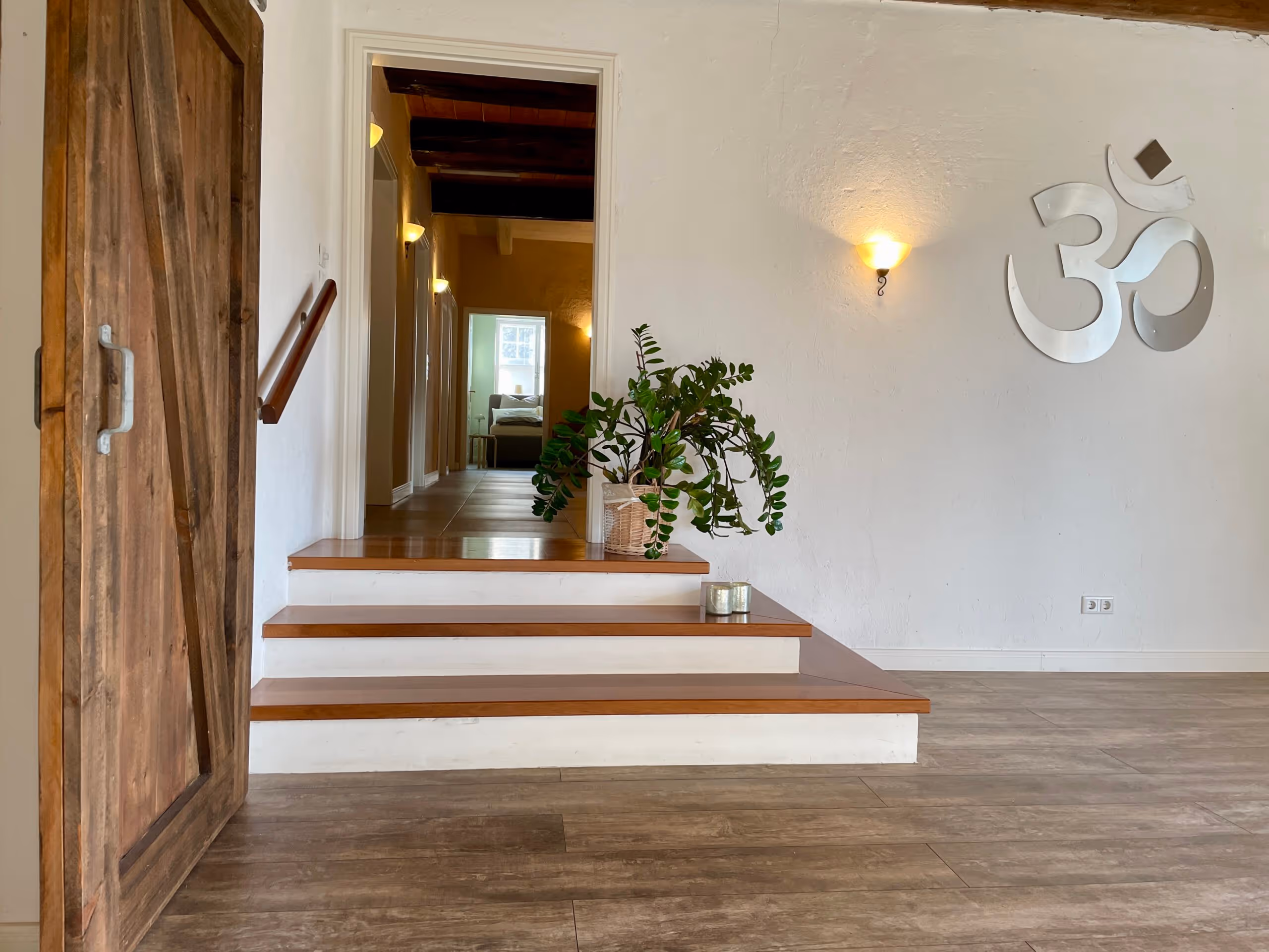 Flur mit Holztür links, drei Holzstufen mit Pflanze auf der obersten Stufe, weißer Wand mit beleuchtetem Om-Symbol und Blick in einen Raum mit Bett am Ende des Gangs.