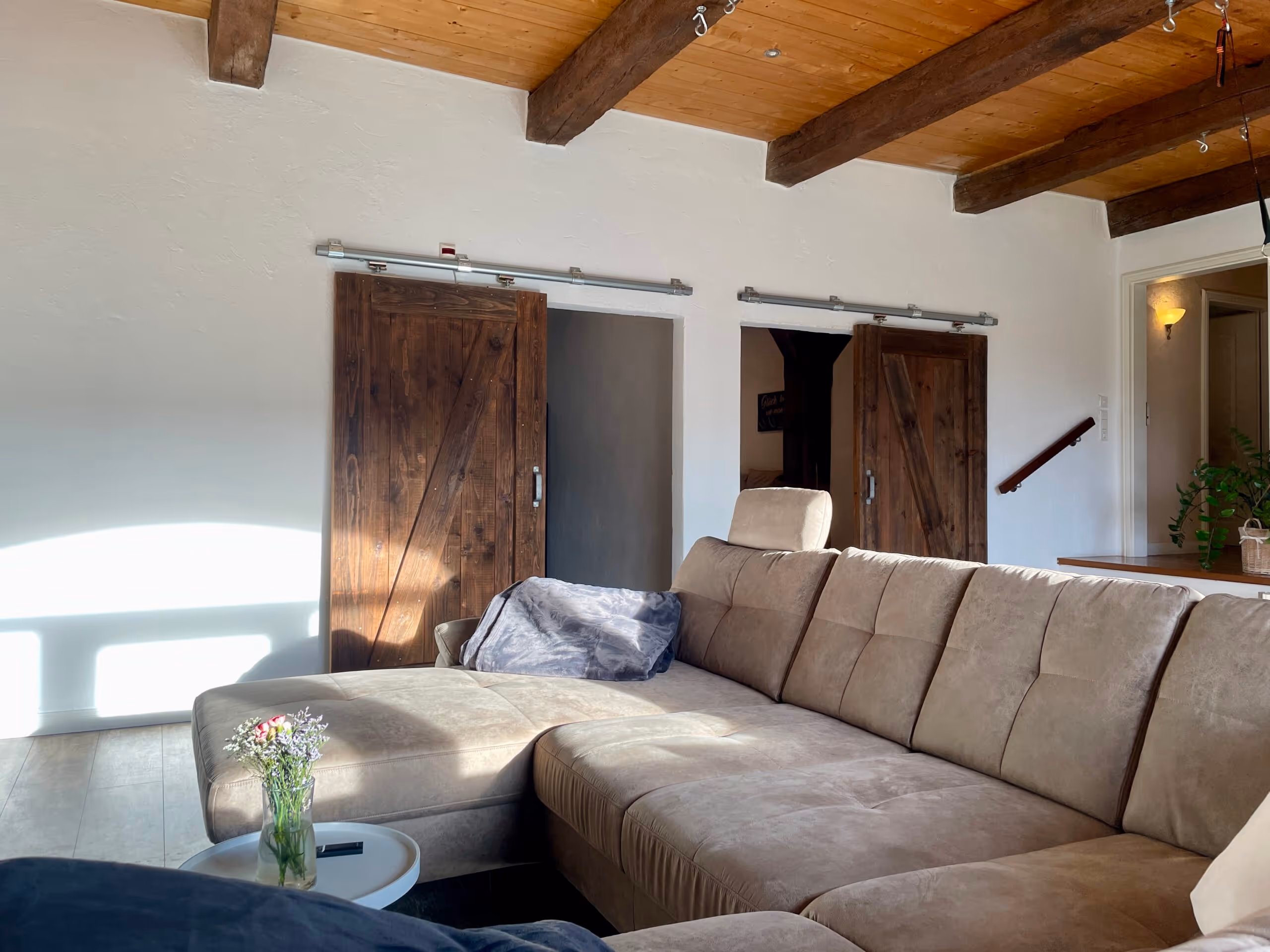 Wohnzimmer mit beigem Ecksofa, kleinen weißen Beistelltisch mit Blumenstrauß, Holzbalkendecke und zwei Schiebetüren aus dunklem Holz.