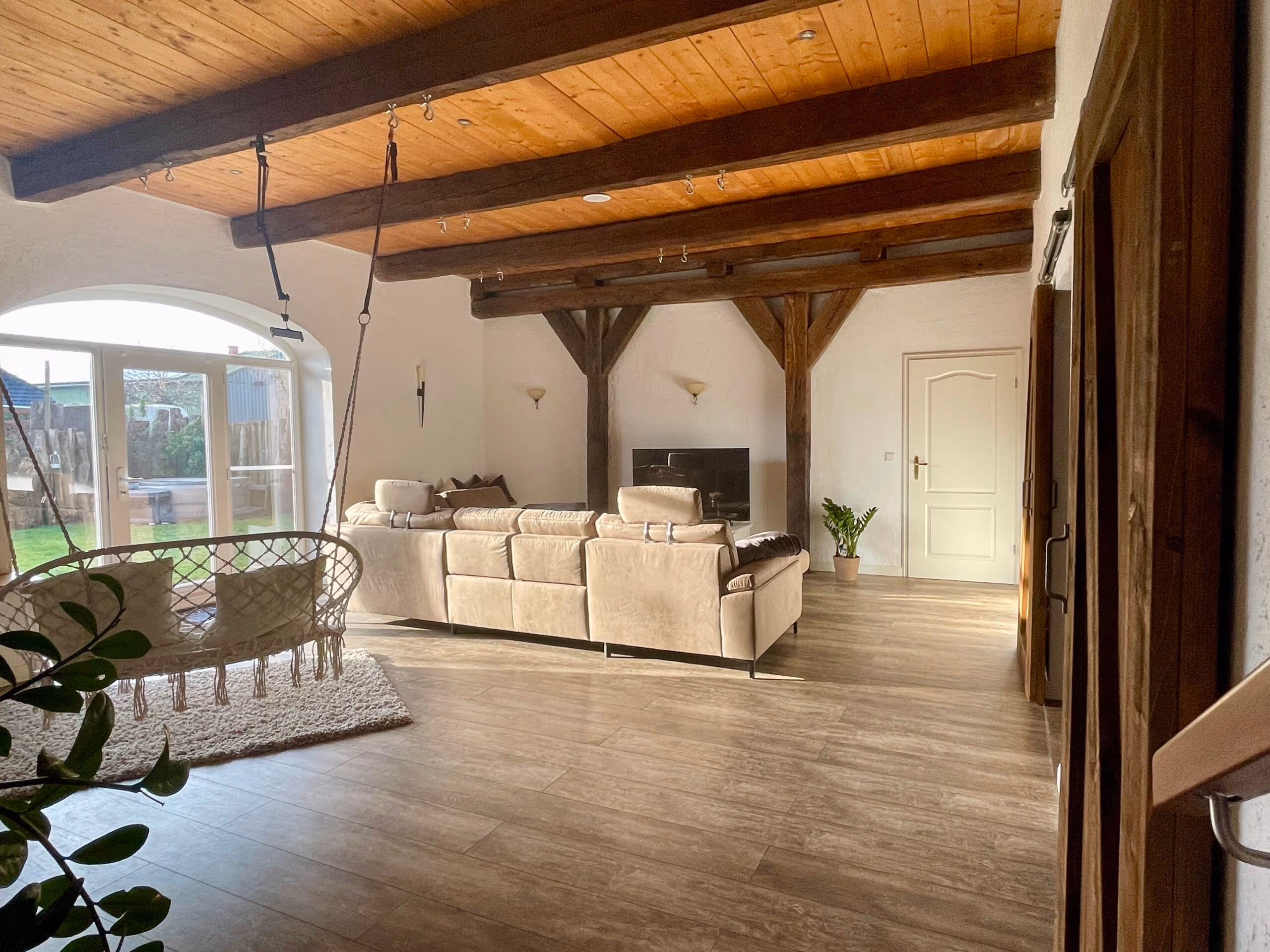 Wohnzimmer mit hellbeiger Ledercouch, hängendem Rattansitz und sichtbaren Holzbalken an Decke.