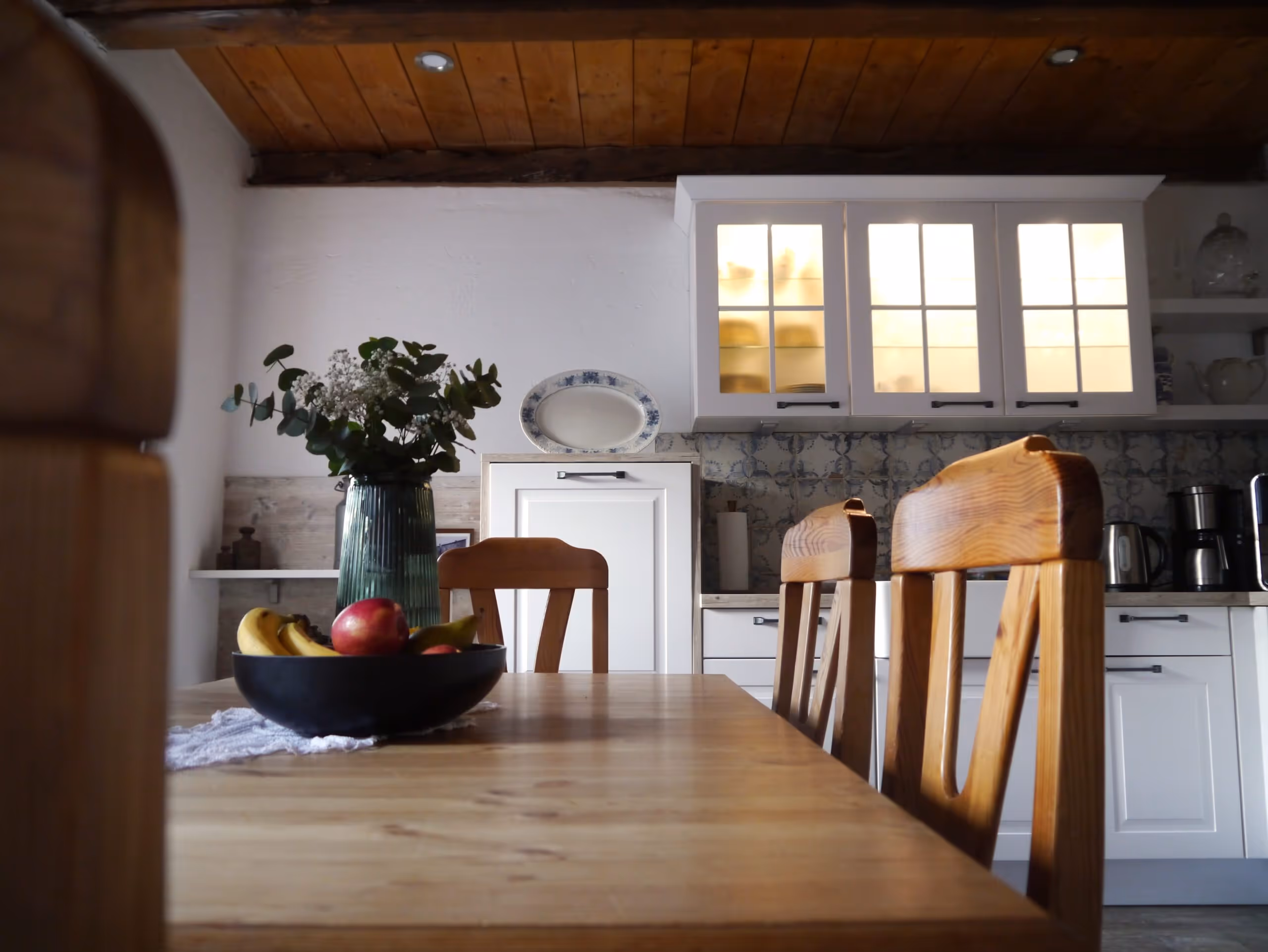 Küche mit Holztisch und Stühlen, Obstschale und Vase mit Blumen, beleuchtete Hängeschränke und Küchenutensilien.