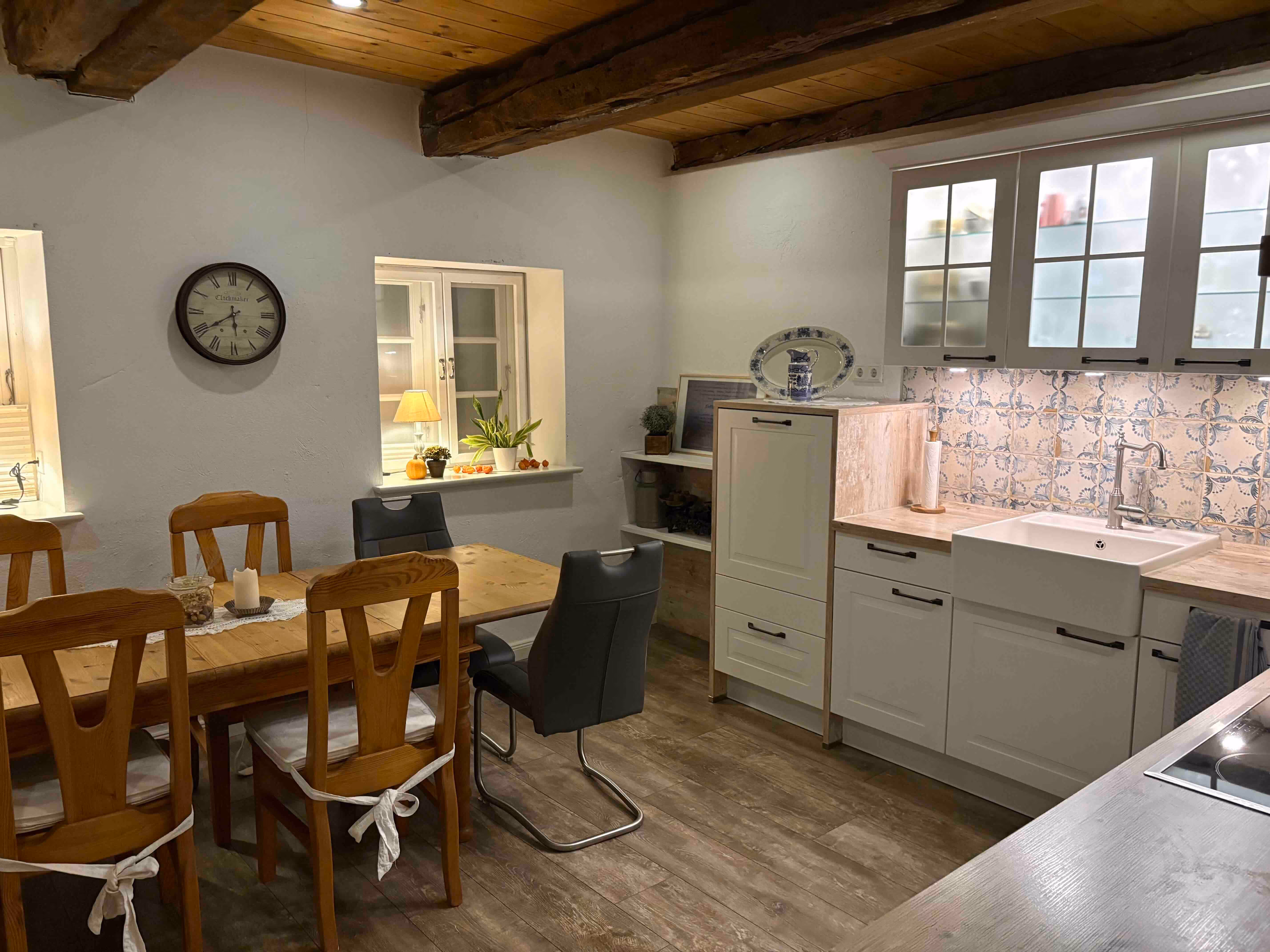 Gemütliche Küche mit Holzbalkendecke, großem Holztisch mit Stühlen, weißen Schränken und Spüle, dekoriert mit Pflanzen und Uhr an der Wand.