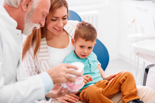 Child and mother having a checkup at dentist office Child having a regular checkup at dentist office, sitting at mother's lap while senior male dentist is demonstrating procedure on plastic jaw model Pediatric dentist in a bright, kid-friendly operatory speaking with a parent and little boy while pointing to a model of the palate stock pictures, royalty-free photos & images