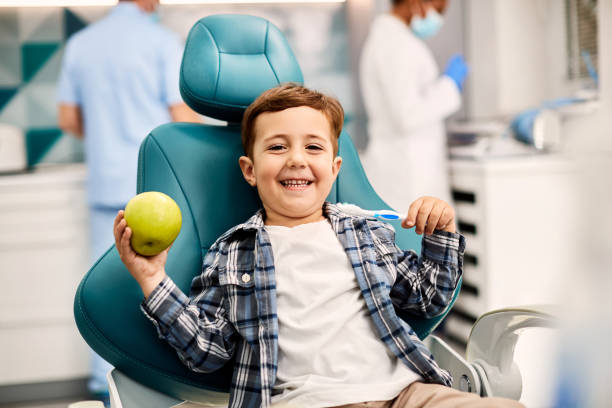 Happy kid holding apple and toothbrush at dentist's office. Happy little boy holding toothbrush and apple while sitting in dentist's chair and looking at camera. A young boy sitting in a bright dental chair, holding a small pumpkin while smiling with the dentist stock pictures, royalty-free photos & images