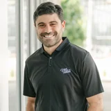 Smiling man with short dark hair and beard wearing a black polo shirt with a logo, standing indoors near a window.