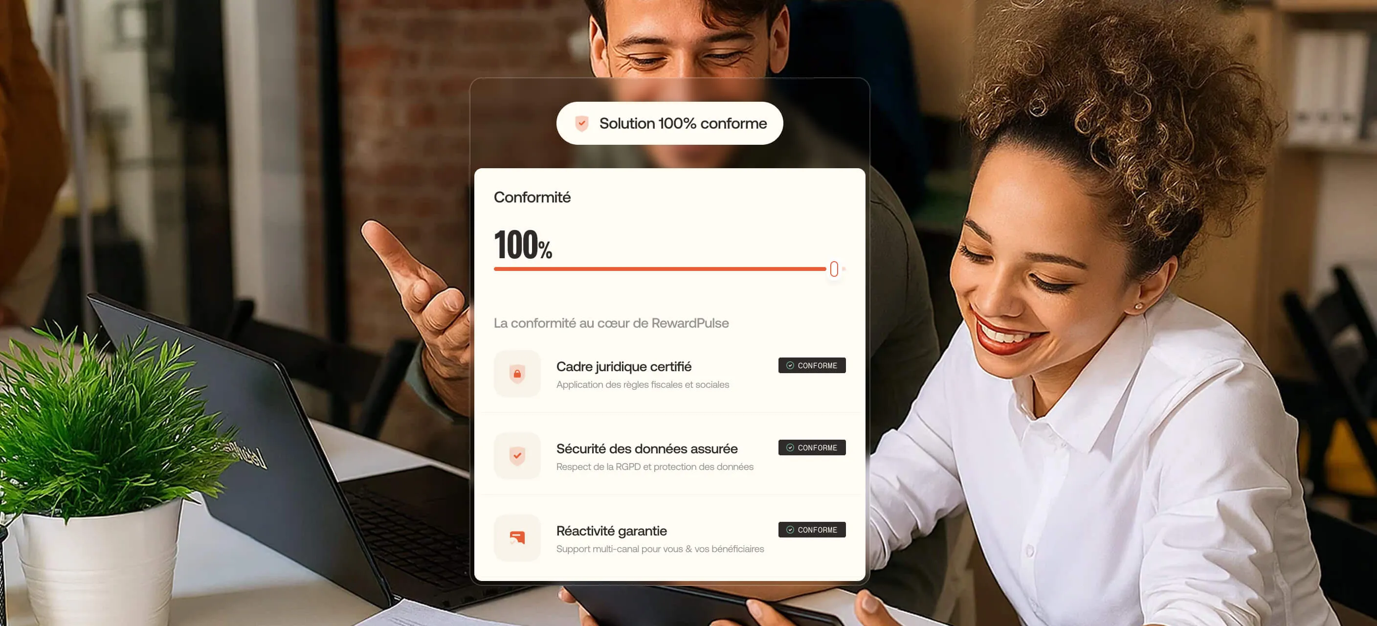 Two colleagues smiling and reviewing information on a tablet in an office setting with a laptop and a potted plant nearby.