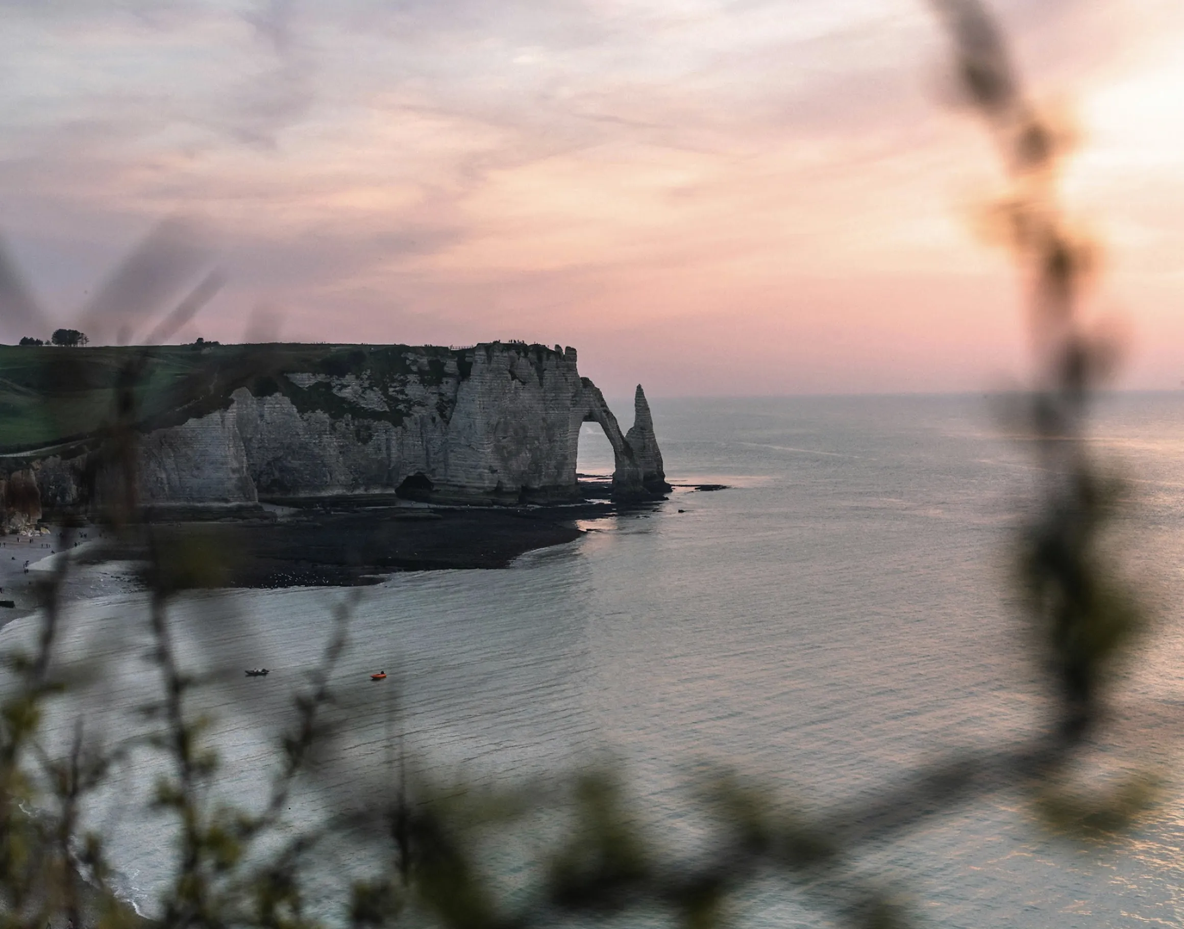 Cliffs with a natural arch formation by a calm sea at sunset, viewed through blurred branches.