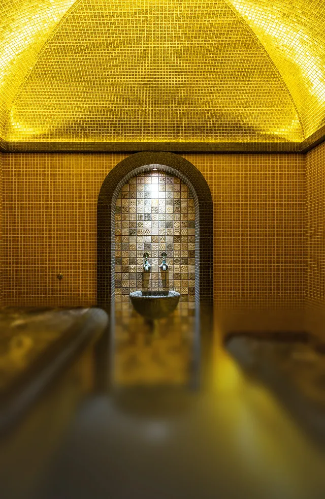 Warmly lit indoor thermal spa with tiled walls and ceiling, featuring a stone basin with two taps under an arched alcove.