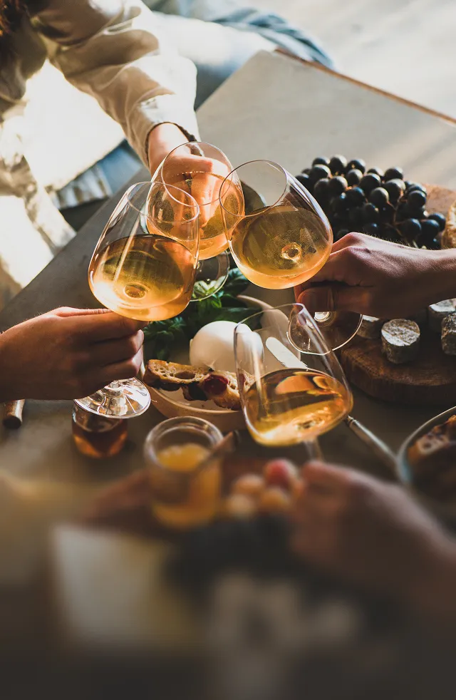 Four people clinking glasses of white wine over a table with grapes, cheese, and bread.