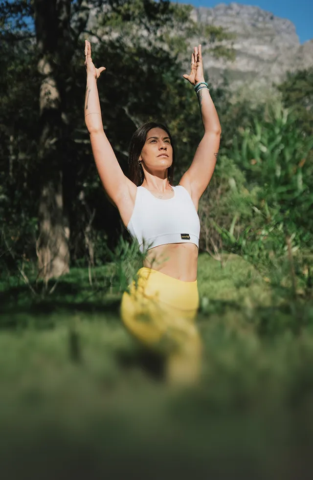 Woman practicing yoga outdoors raising arms upwards with a focused expression, surrounded by greenery and mountains in the background.