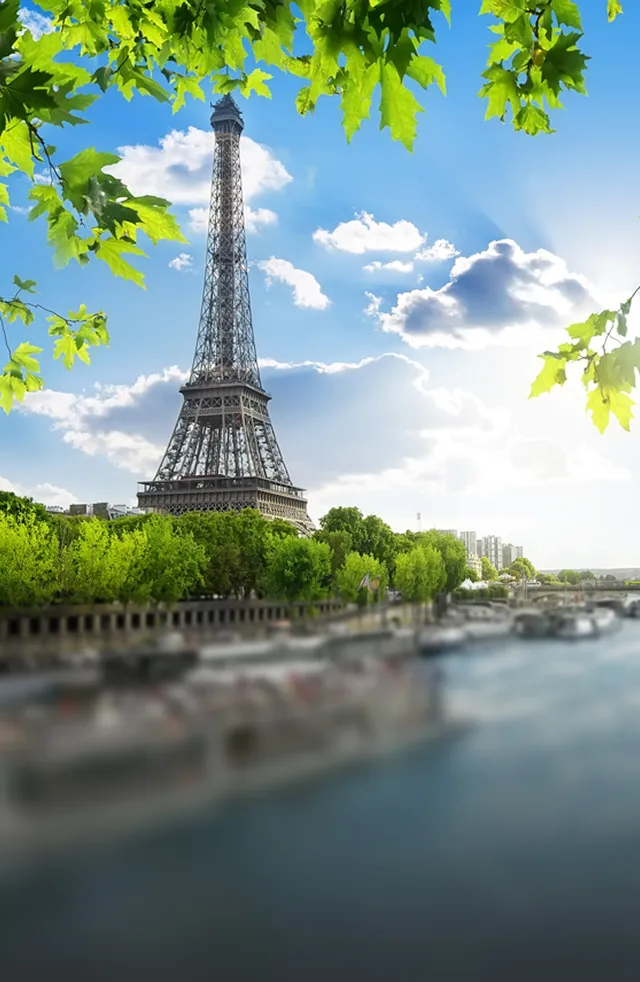 Eiffel Tower in Paris framed by green leaves with the Seine River and cloudy blue sky in the background.