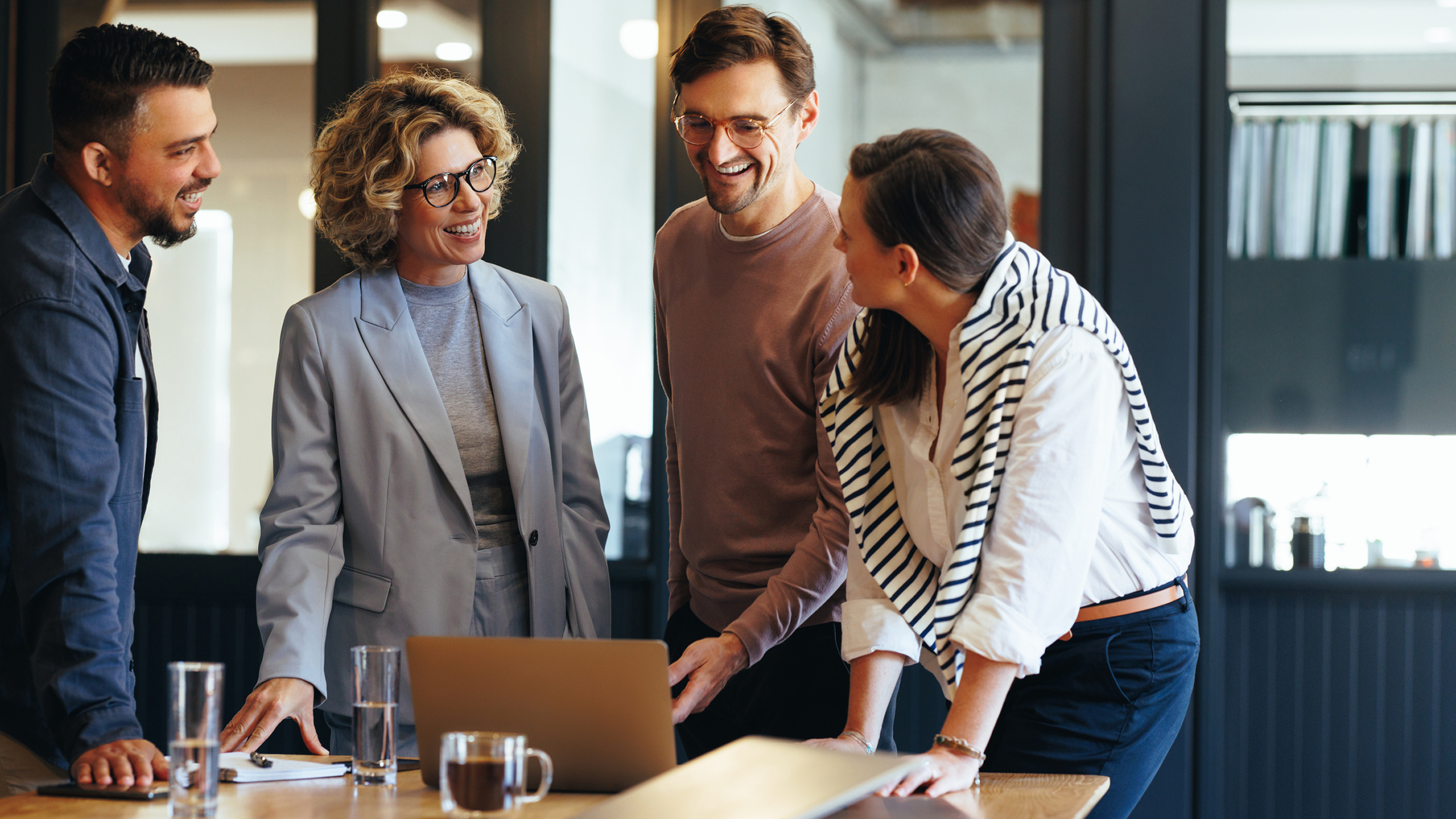 Four colleagues smiling and discussing around a laptop in a modern office.