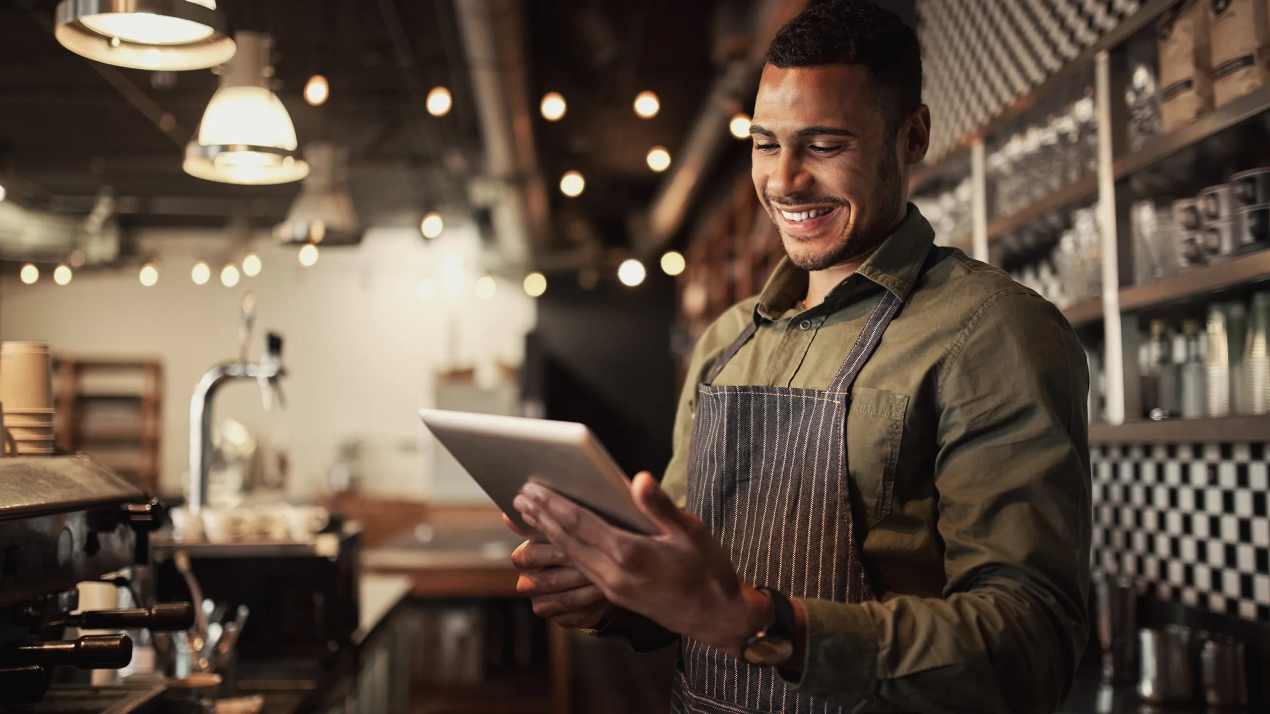 Smiling barista wearing an apron holding a tablet inside a modern coffee shop.