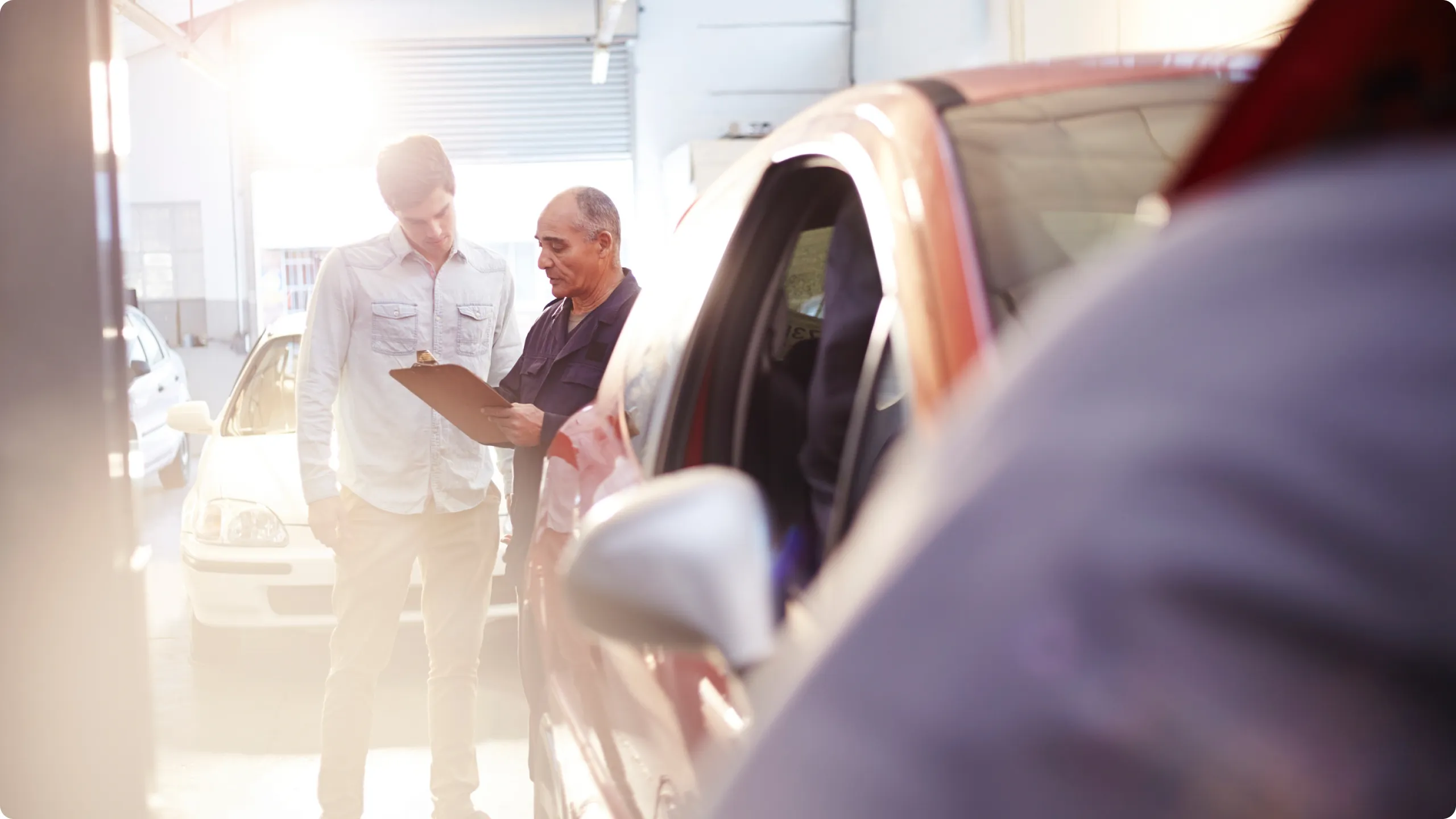 Two men standing indoors near a red car, one holding a clipboard while they discuss.