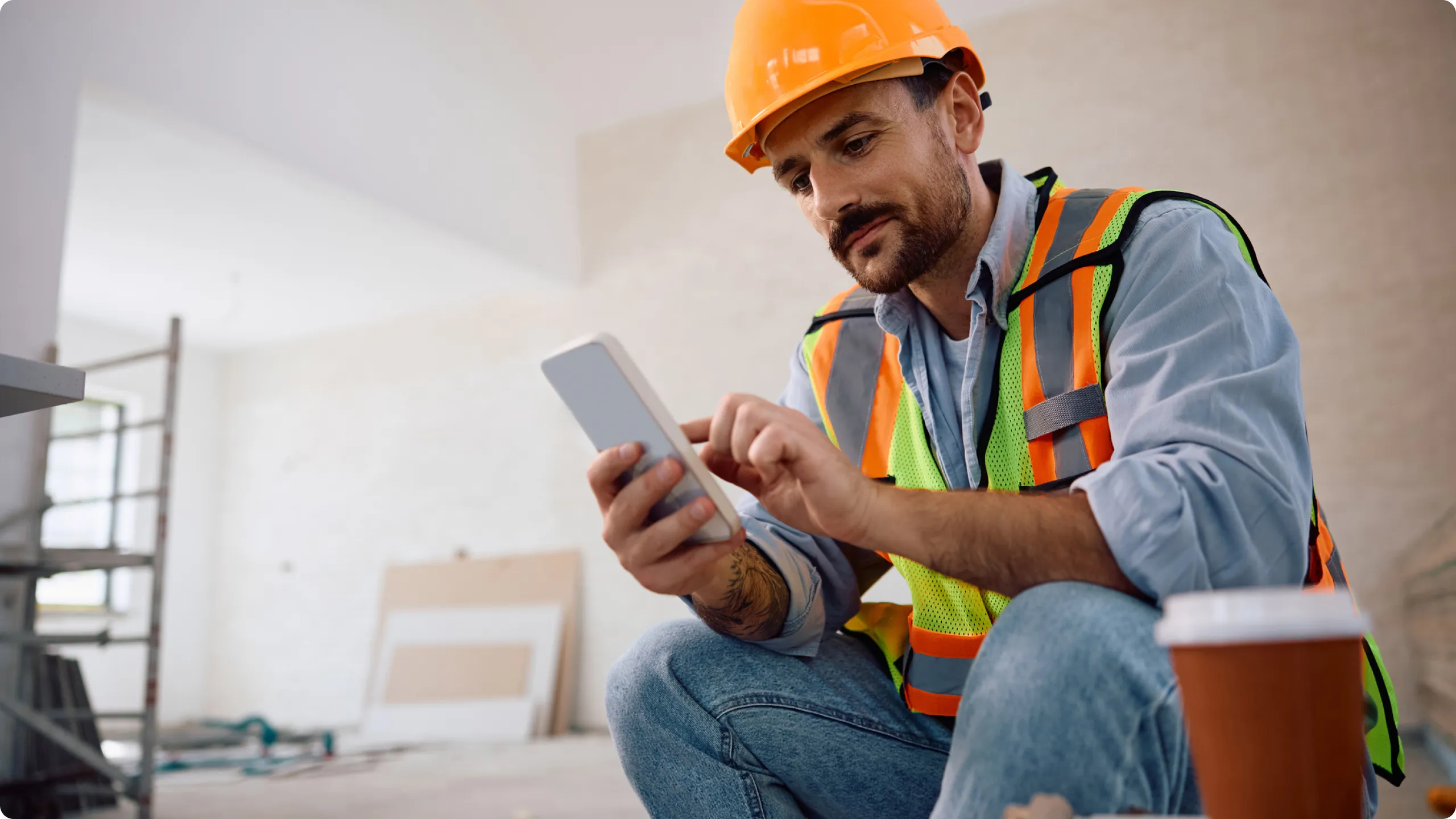 Construction worker wearing an orange hard hat and safety vest using a smartphone indoor on a construction site.
