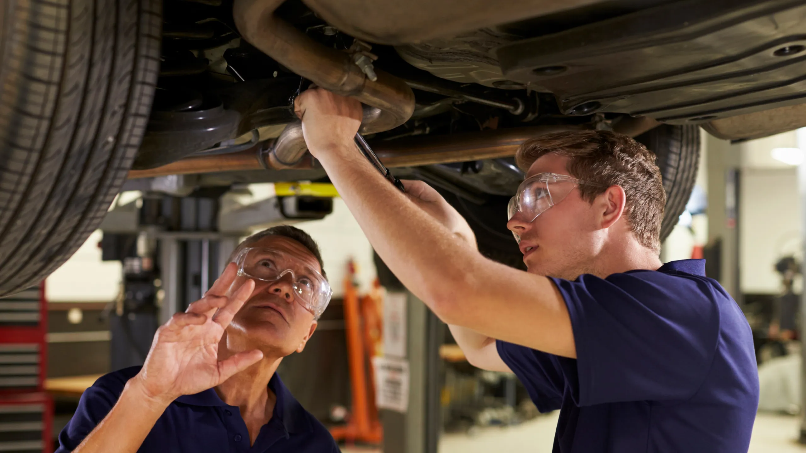 Two mechanics wearing safety glasses working under a raised car, one using a tool and the other observing.