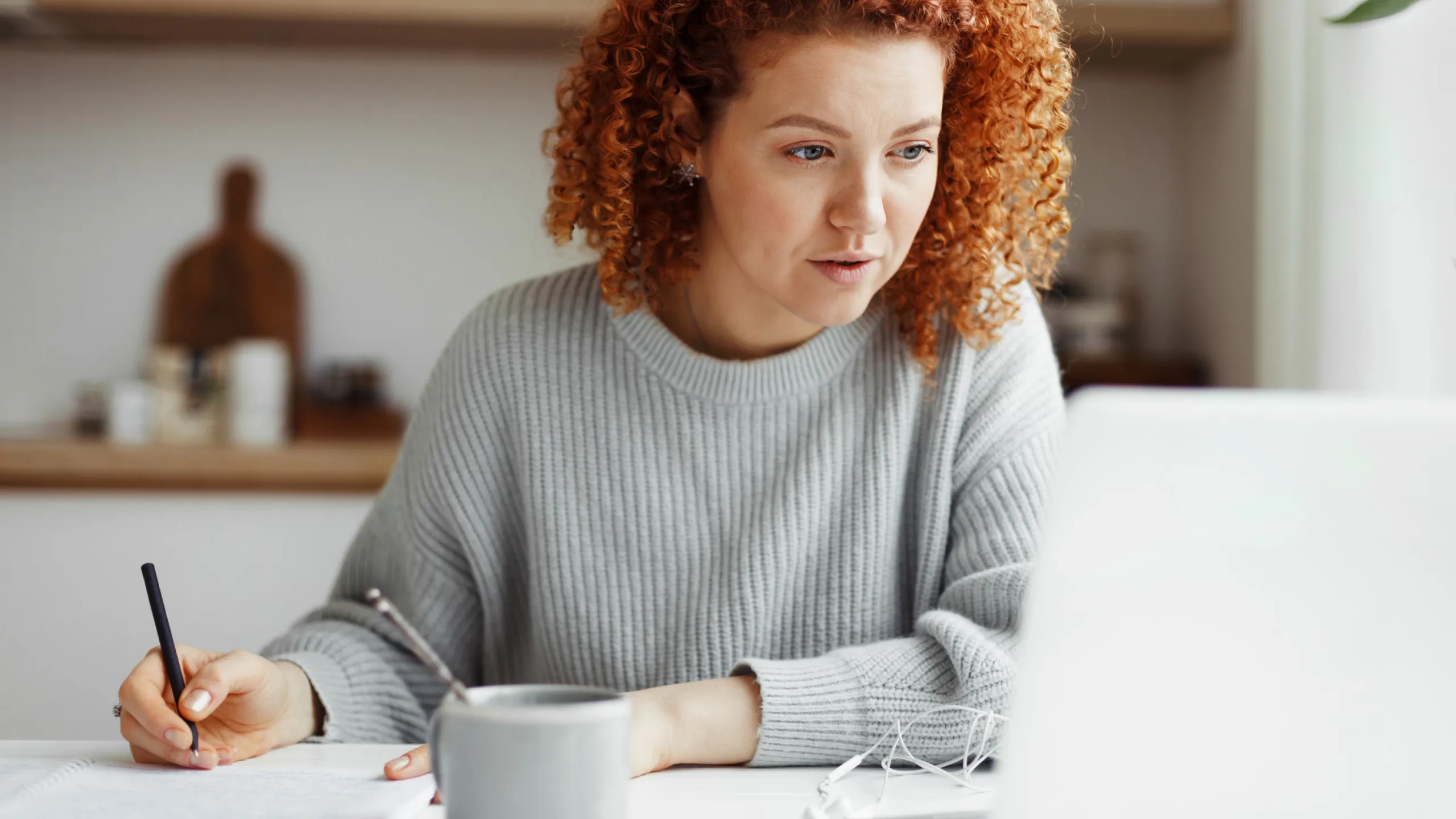 Woman with curly red hair focusing on her laptop while writing in a notebook at a table.