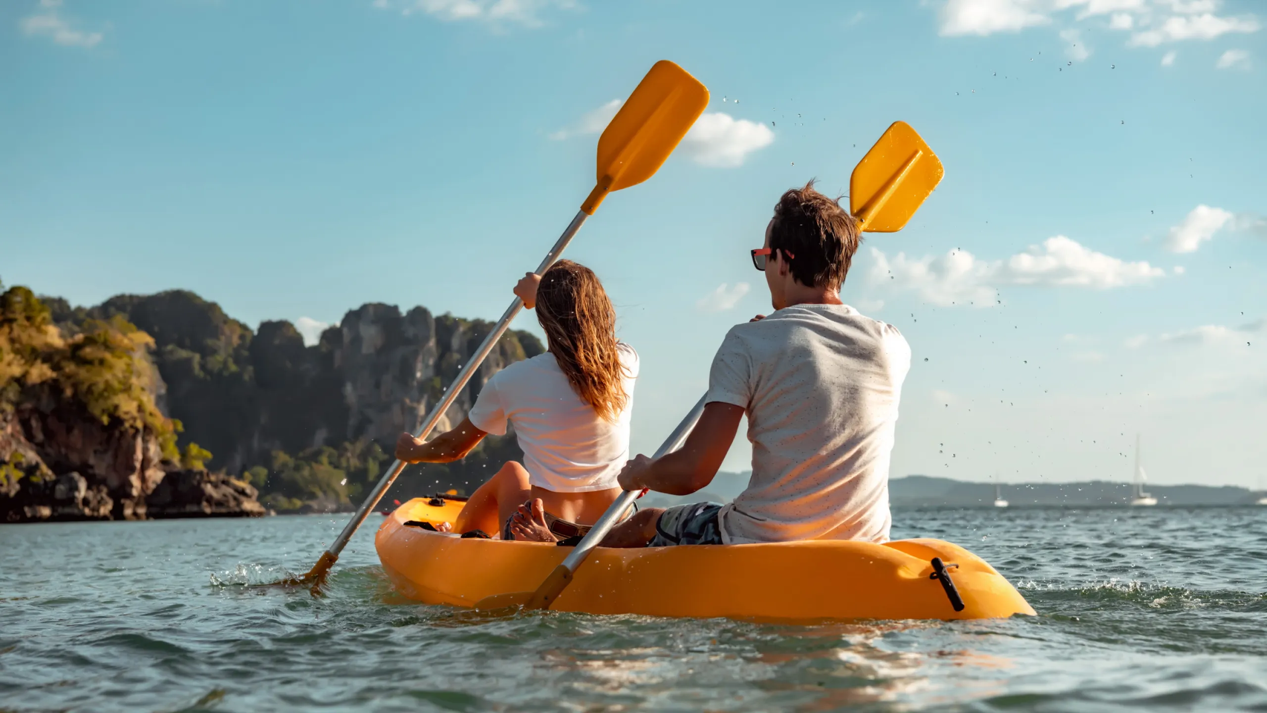 Man and woman paddling a yellow kayak on a calm body of water with rocky cliffs and a partly cloudy sky in the background.