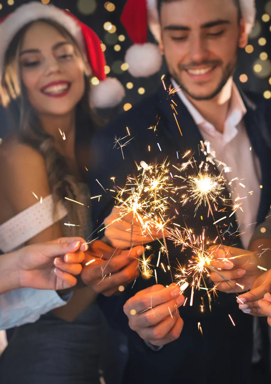 Smiling people wearing Santa hats holding sparklers with festive bokeh lights in the background.