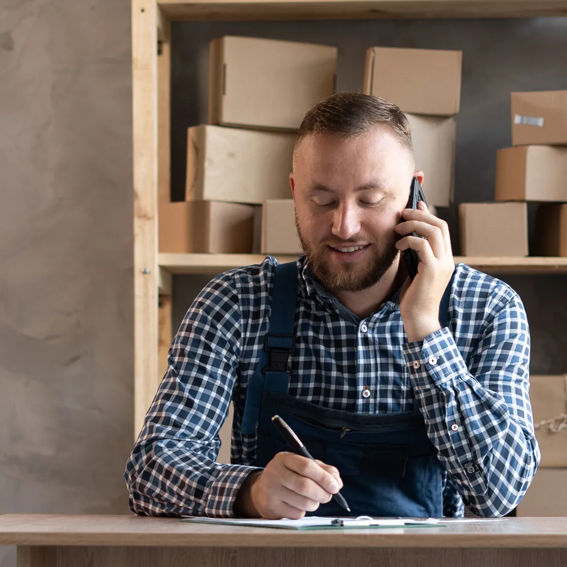Man in plaid shirt and overalls talking on phone and writing on clipboard at table with stacked cardboard boxes behind him.