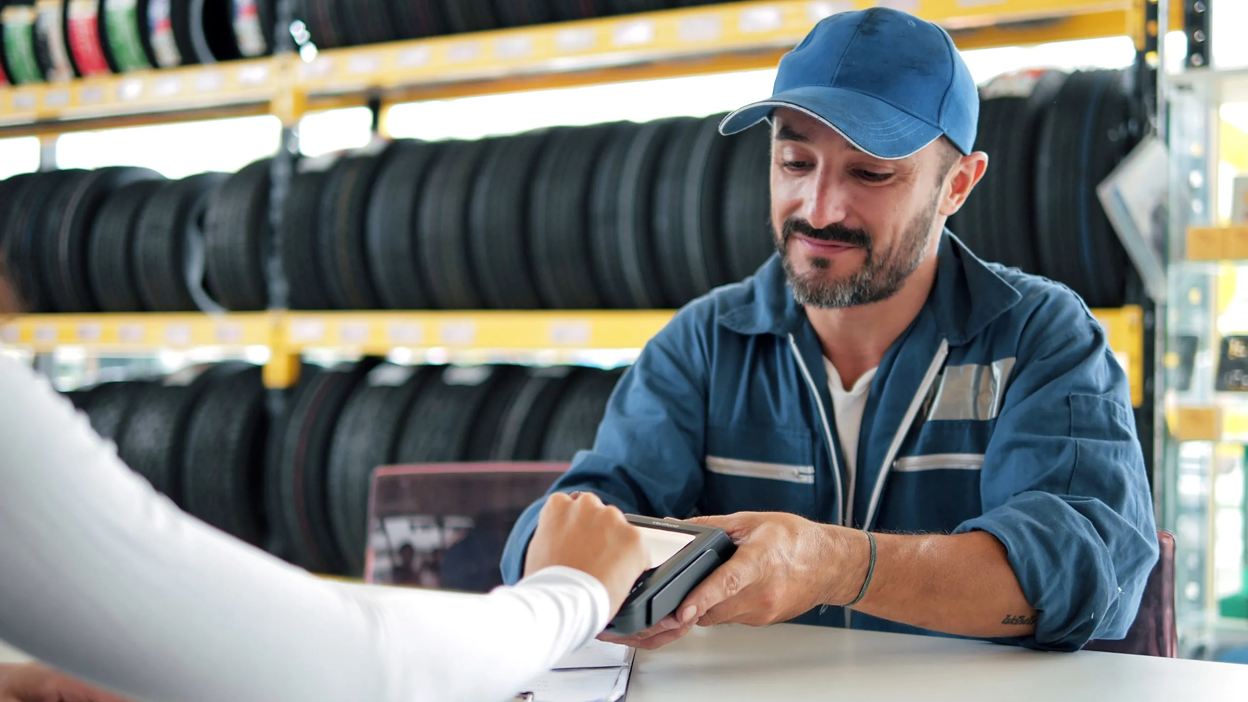 Man in blue work uniform and cap holding a card payment terminal as a customer makes a payment inside a tire shop.