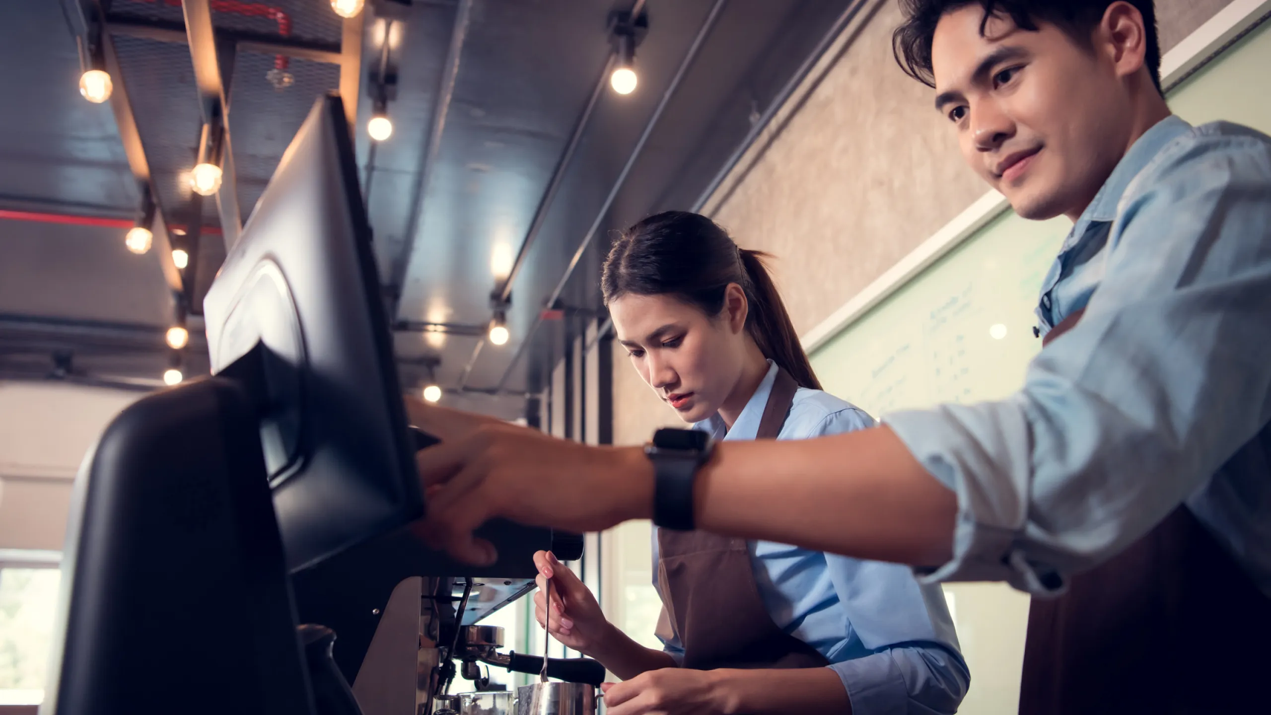 Two baristas in aprons working together on a coffee machine in a cafe.