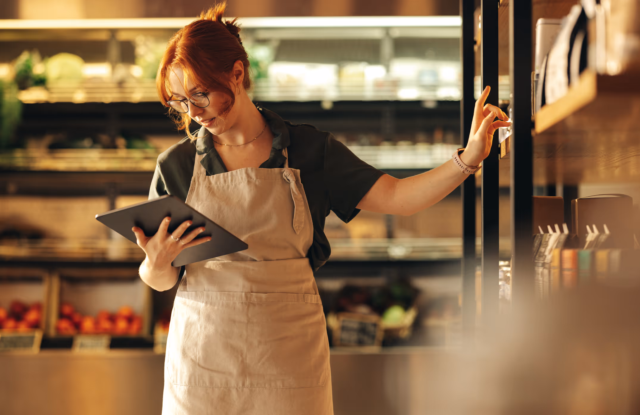 Woman wearing apron and glasses holding a tablet while organizing items on a store shelf.