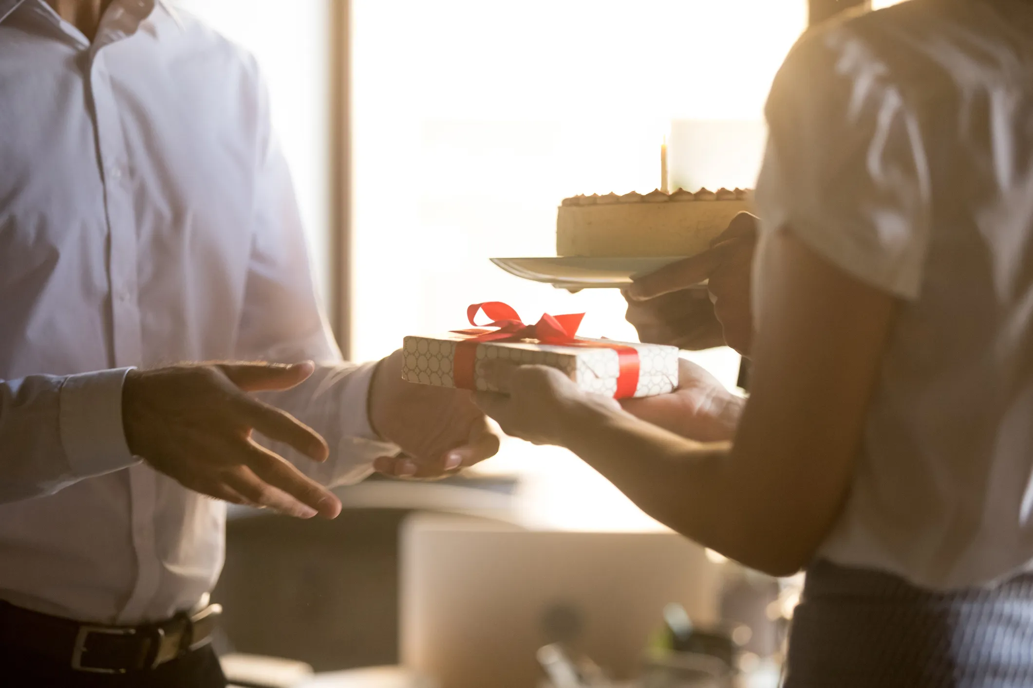 Two people exchanging a birthday cake with a lit candle and a gift wrapped with a red ribbon in warm sunlight.