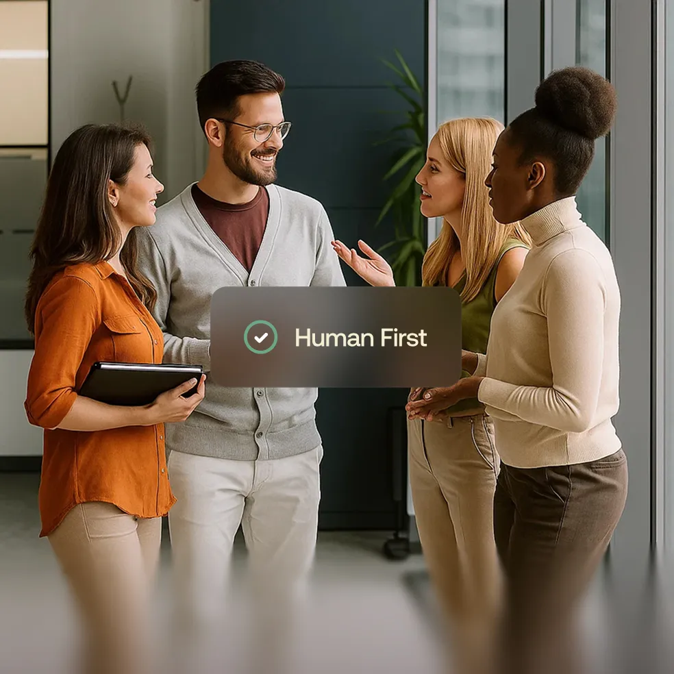 Four diverse colleagues standing and having a friendly discussion in an office with a 'Human First' checkmark overlay.