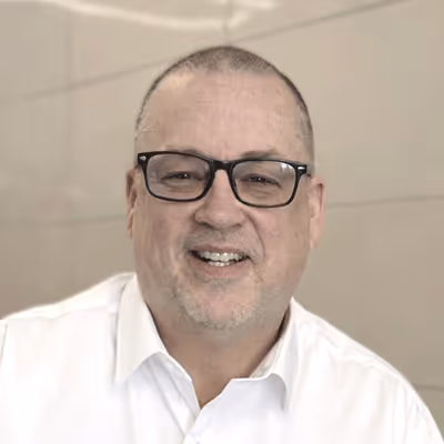 Smiling middle-aged man with short hair and glasses wearing a white shirt against a neutral background.
