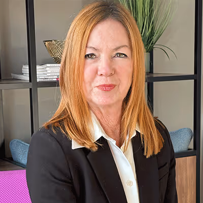 Woman with straight red hair wearing a black blazer and white collared shirt, seated in an office with shelves and plants behind her.