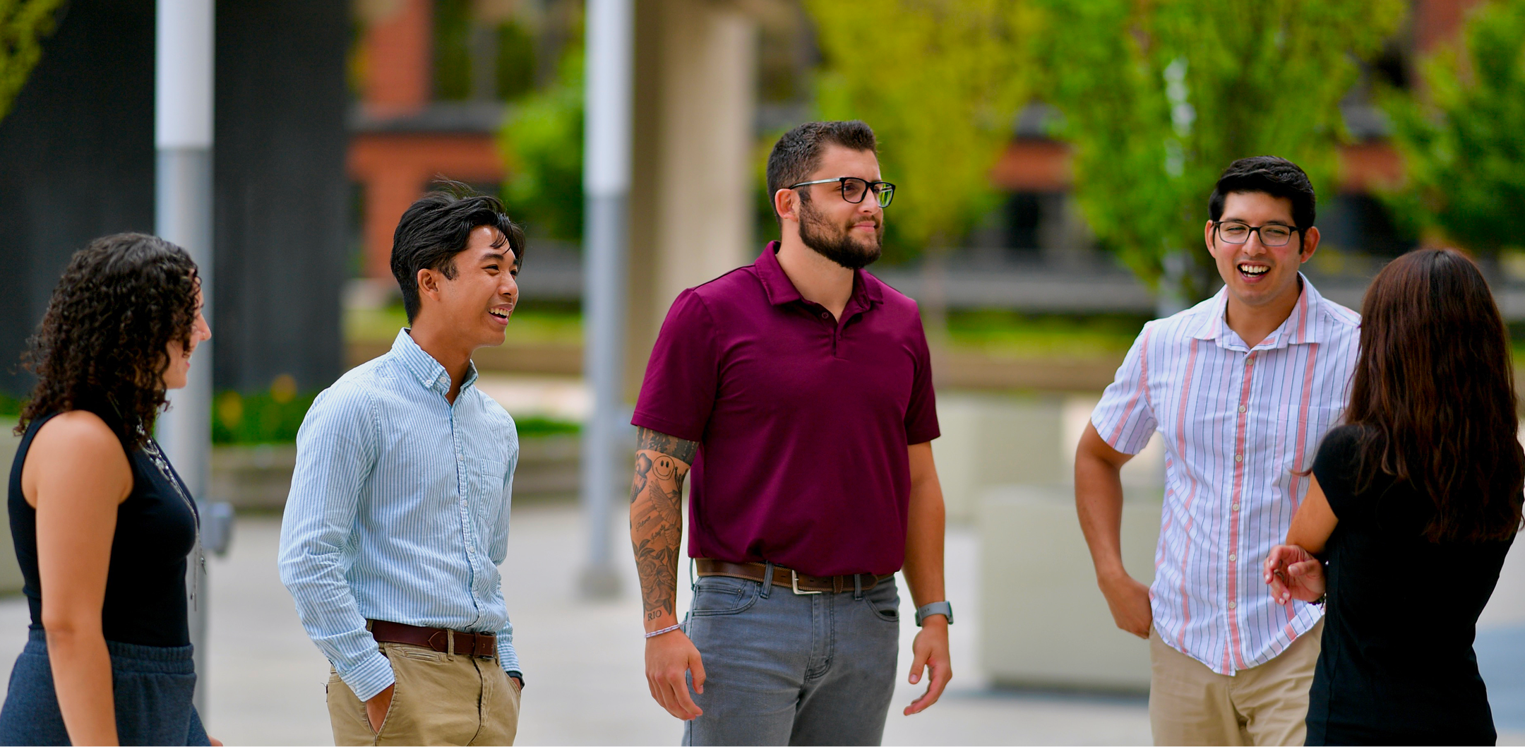 Five diverse young adults standing outdoors in a casual conversation with greenery and buildings in the background.
