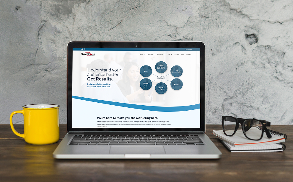 Open laptop displaying a marketing website with text about understanding audience and marketing solutions, yellow mug on left, black glasses and notebooks on right on wooden table.