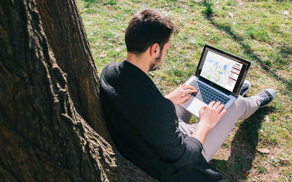 Man sitting on grass under a tree working on a laptop displaying a digital map.