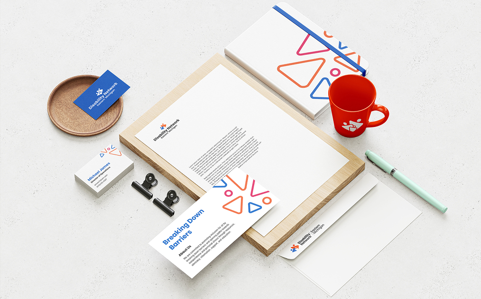 Desk layout with branded stationery including letterhead, business cards, brochure, notebook, pen, envelope, red mug, and wooden tray with clips.