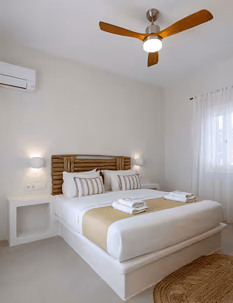 Minimalist bedroom with a bed dressed in white and beige linens, wooden headboard, ceiling fan, and soft natural light from a sheer curtained window.