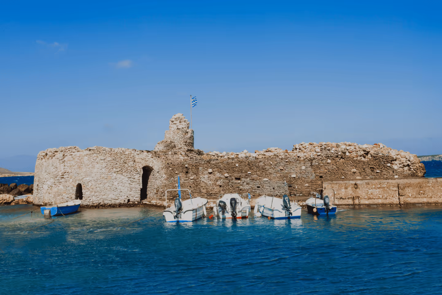 Small motorboats moored in clear blue water next to an ancient stone fort with a Greek flag flying.
