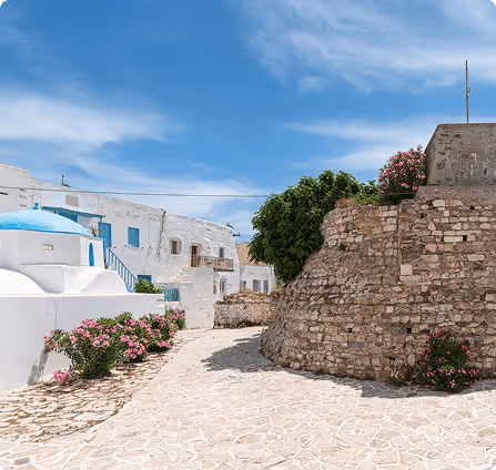 Cobblestone pathway between white buildings with a blue-domed church and stone wall under a blue sky.