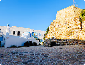 Sunlit stone-paved courtyard beside white Mediterranean buildings and a large stone wall under a clear blue sky.