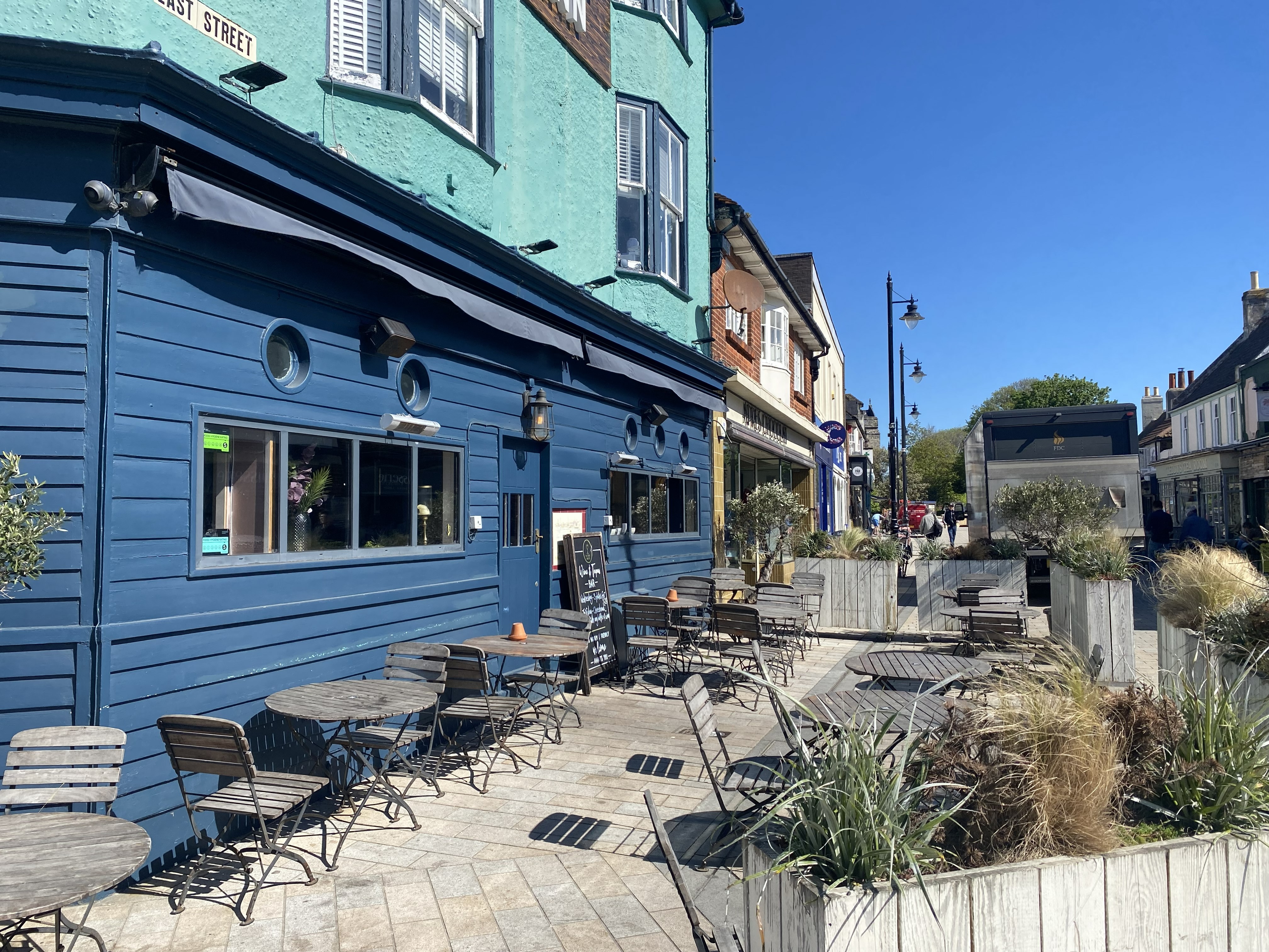 Outdoor seating area with wooden tables and chairs along a blue building on a sunny street.