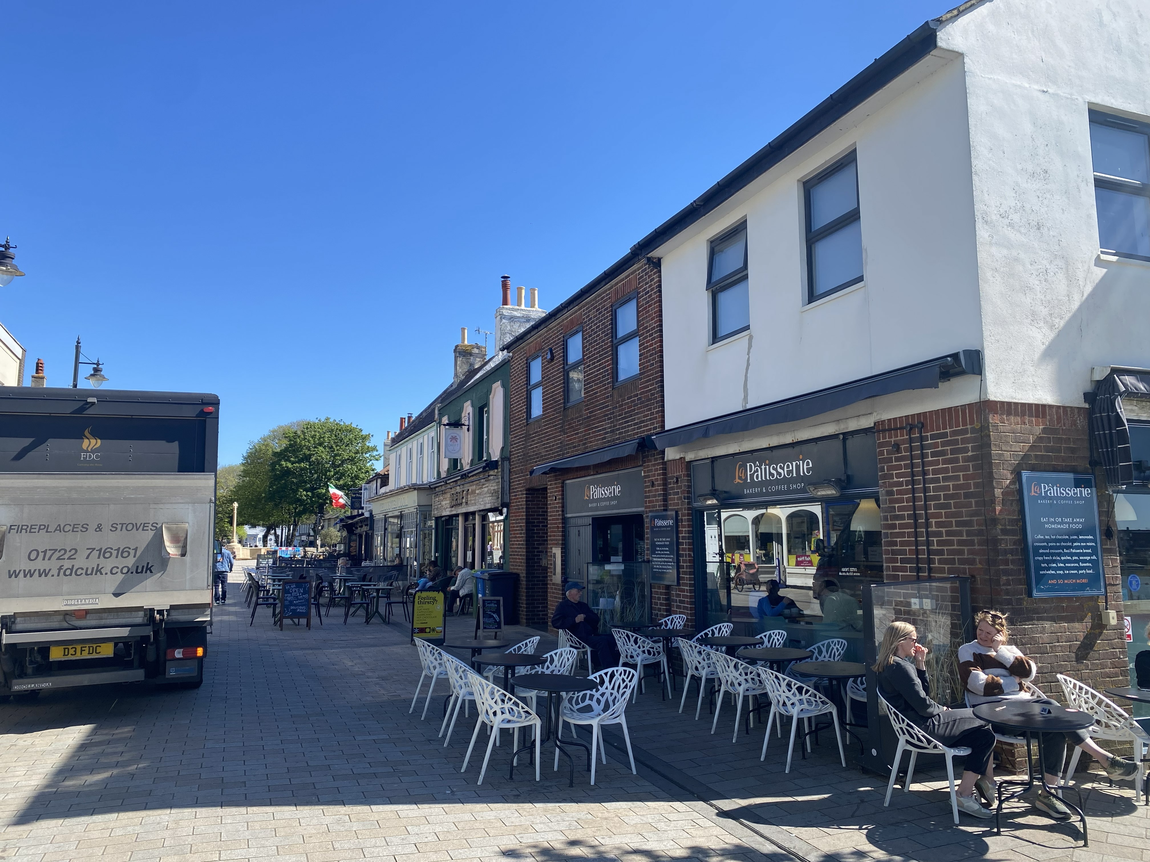 Outdoor seating area of La Pâtisserie bakery and coffee shop on a sunny street with people sitting at tables and a delivery truck parked nearby.