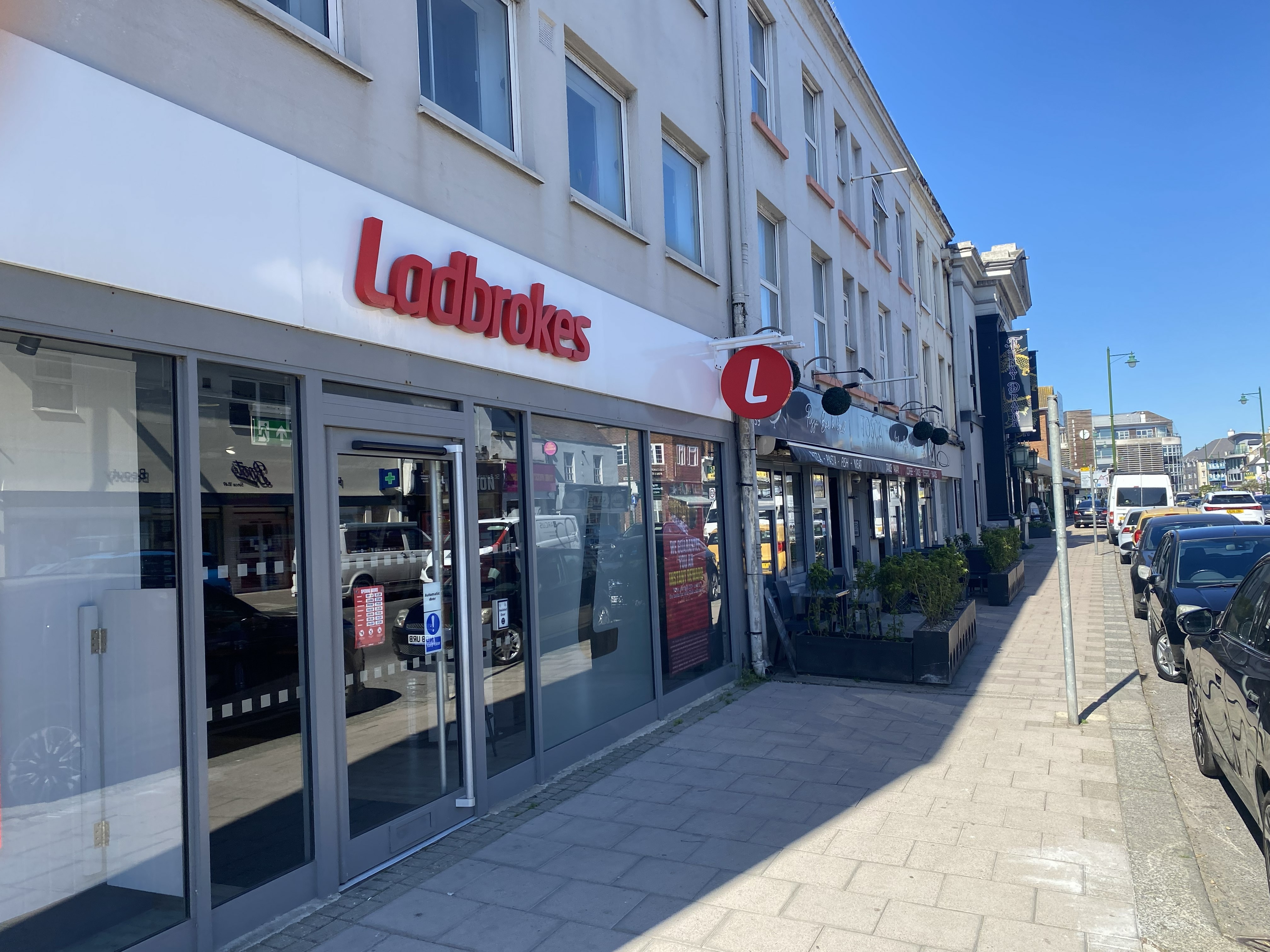 Exterior of a Ladbrokes betting shop on a sunny street with parked cars and neighboring businesses.