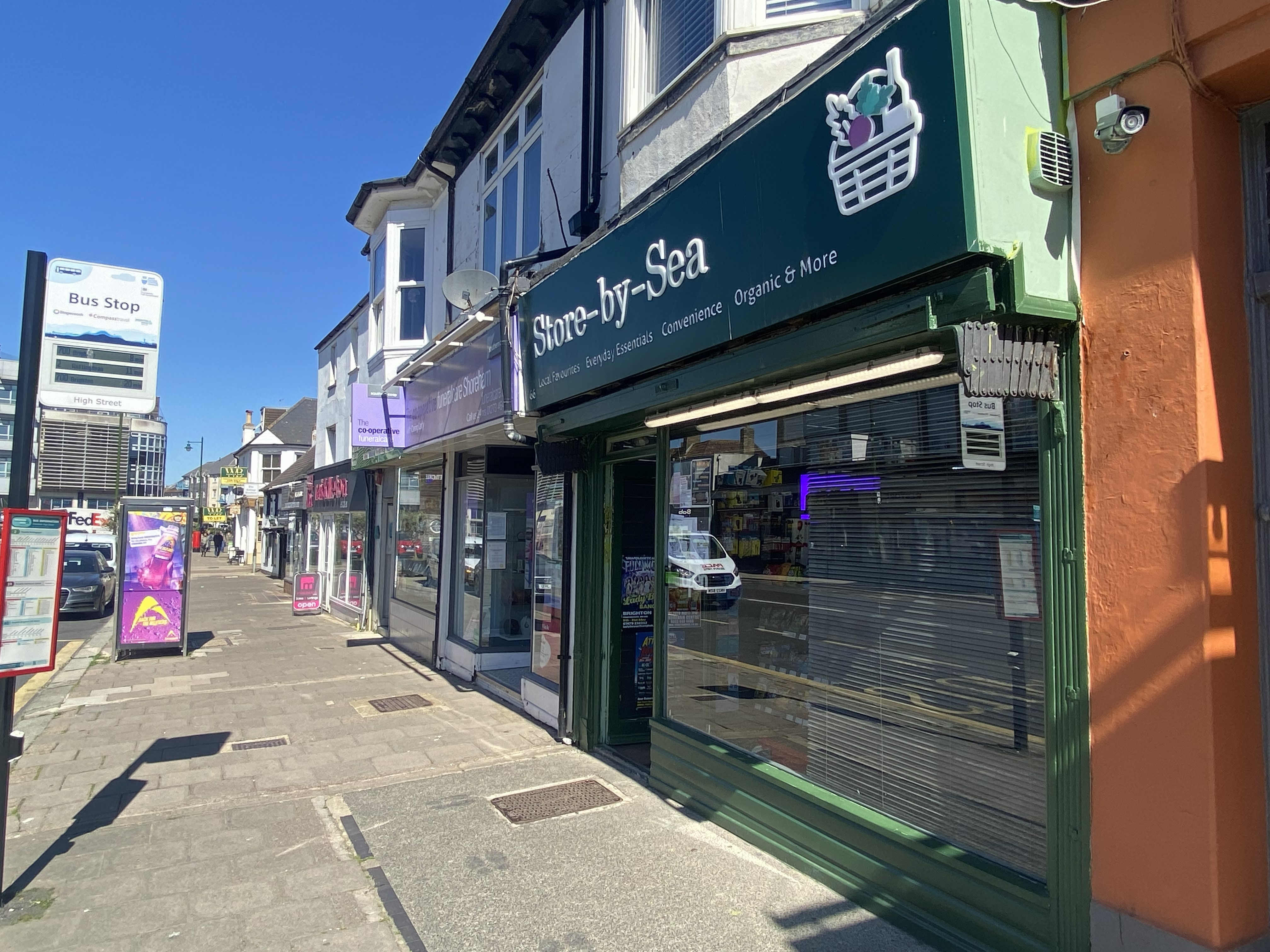 Street view of a row of shops with a green storefront named Store-by-Sea under a clear blue sky and a nearby bus stop sign.