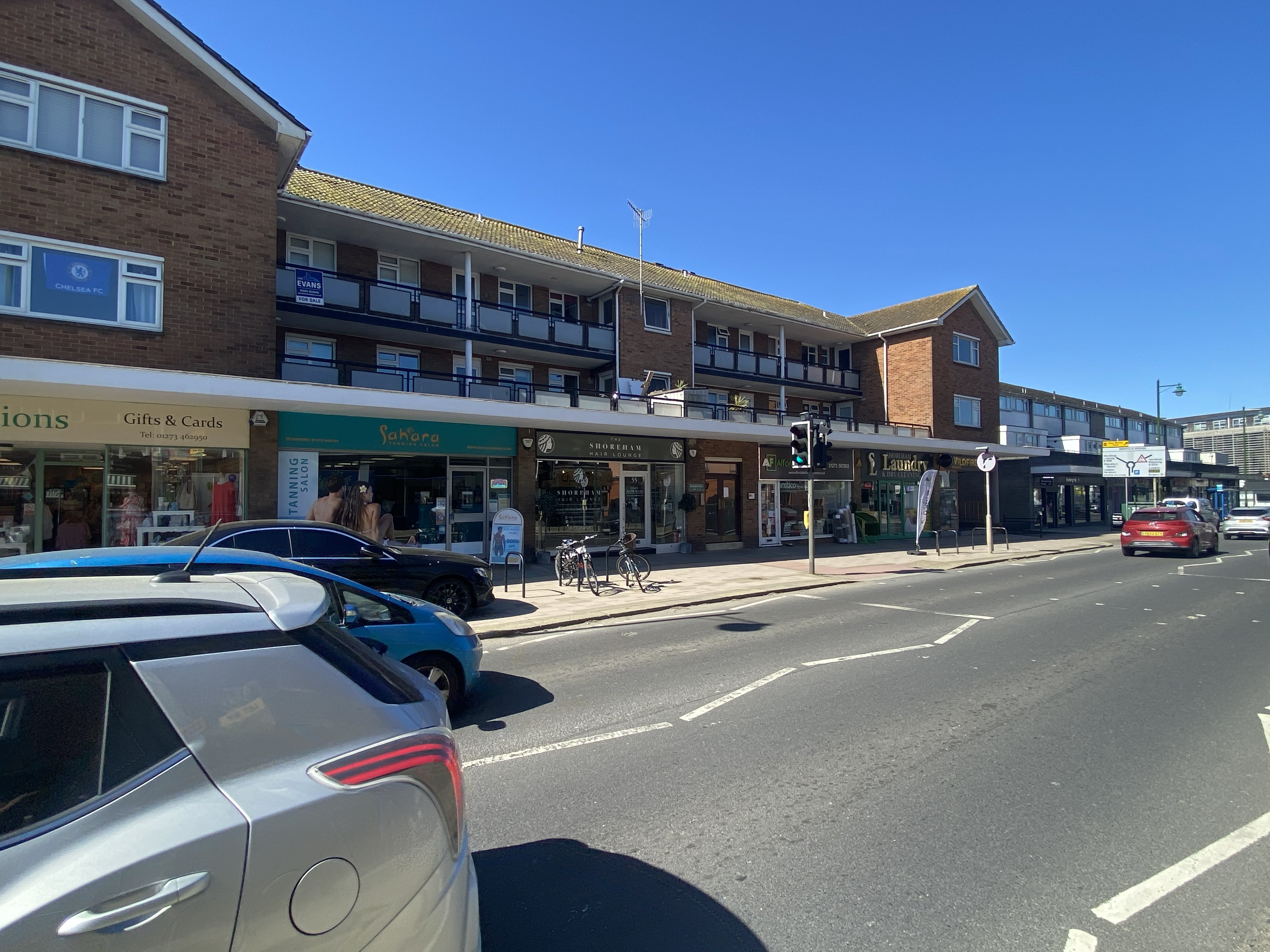 Street view of a commercial building with shops including a tanning salon, hair lounge, and a laundry, under a clear blue sky with parked cars and bicycles.