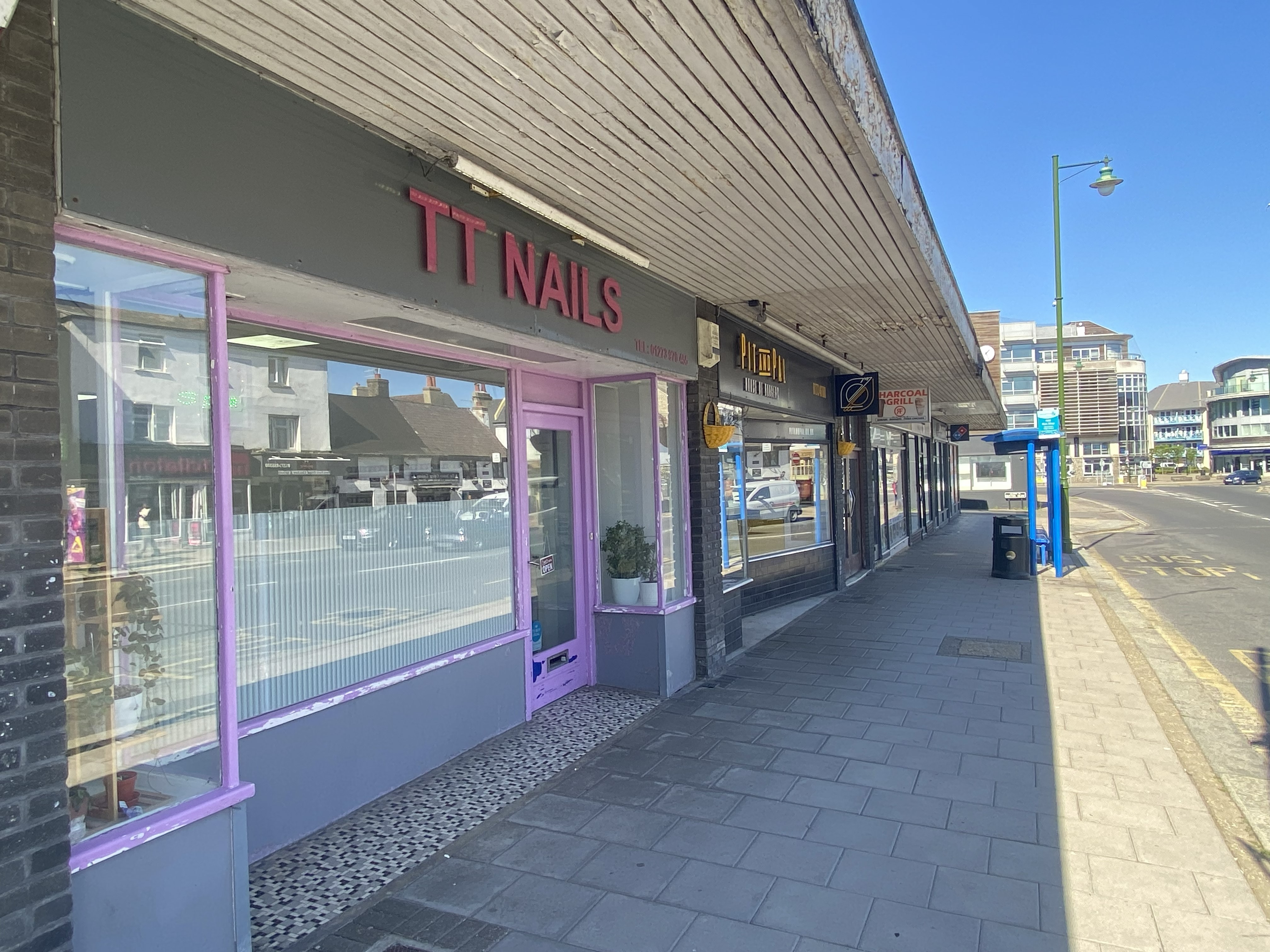 Sidewalk with shops including TT Nails with purple trim and a bus stop on a sunny day.