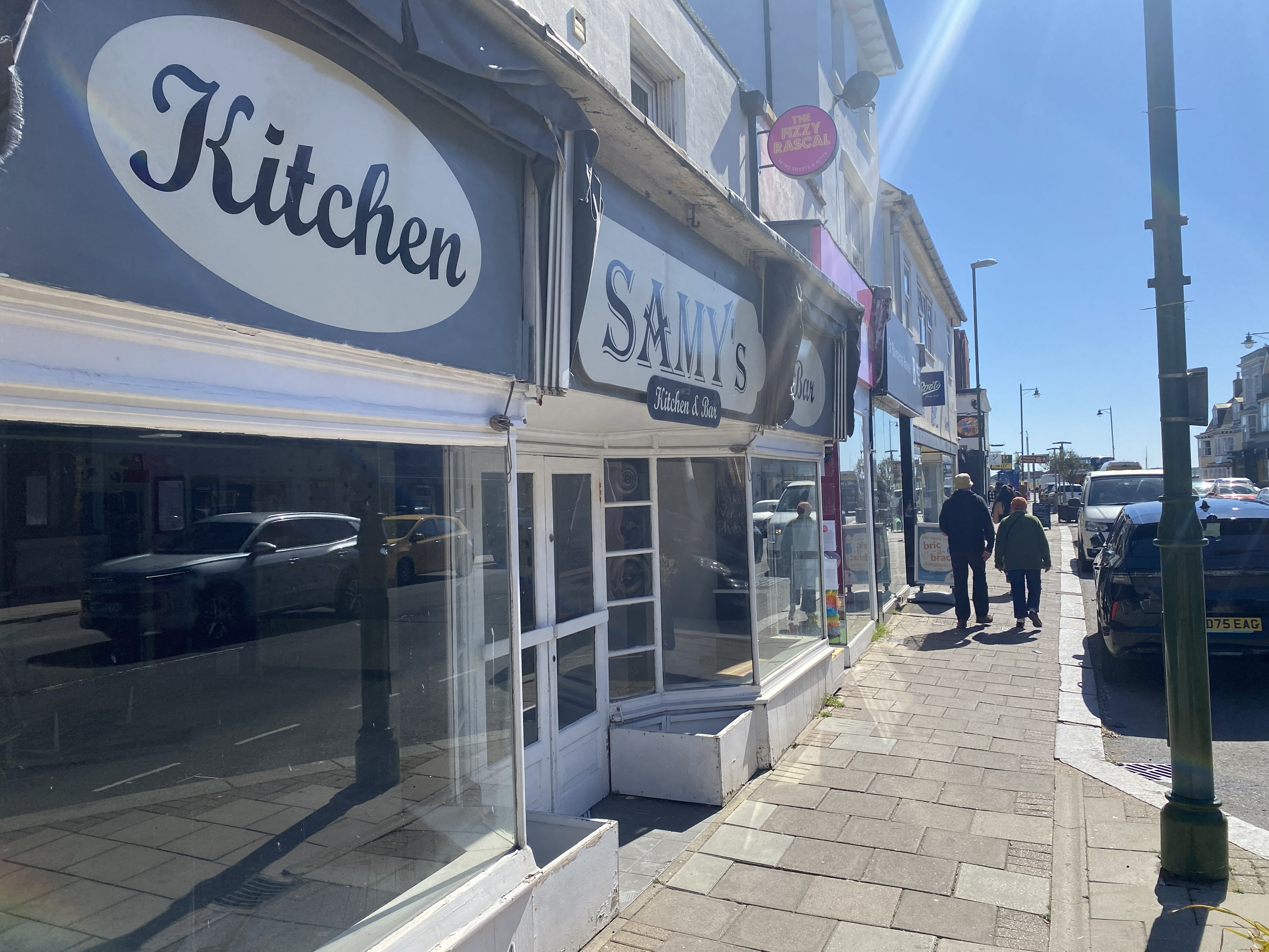 Street view of a white storefront with signs reading 'Kitchen' and 'SAMY's Kitchen & Bar' under a clear blue sky.