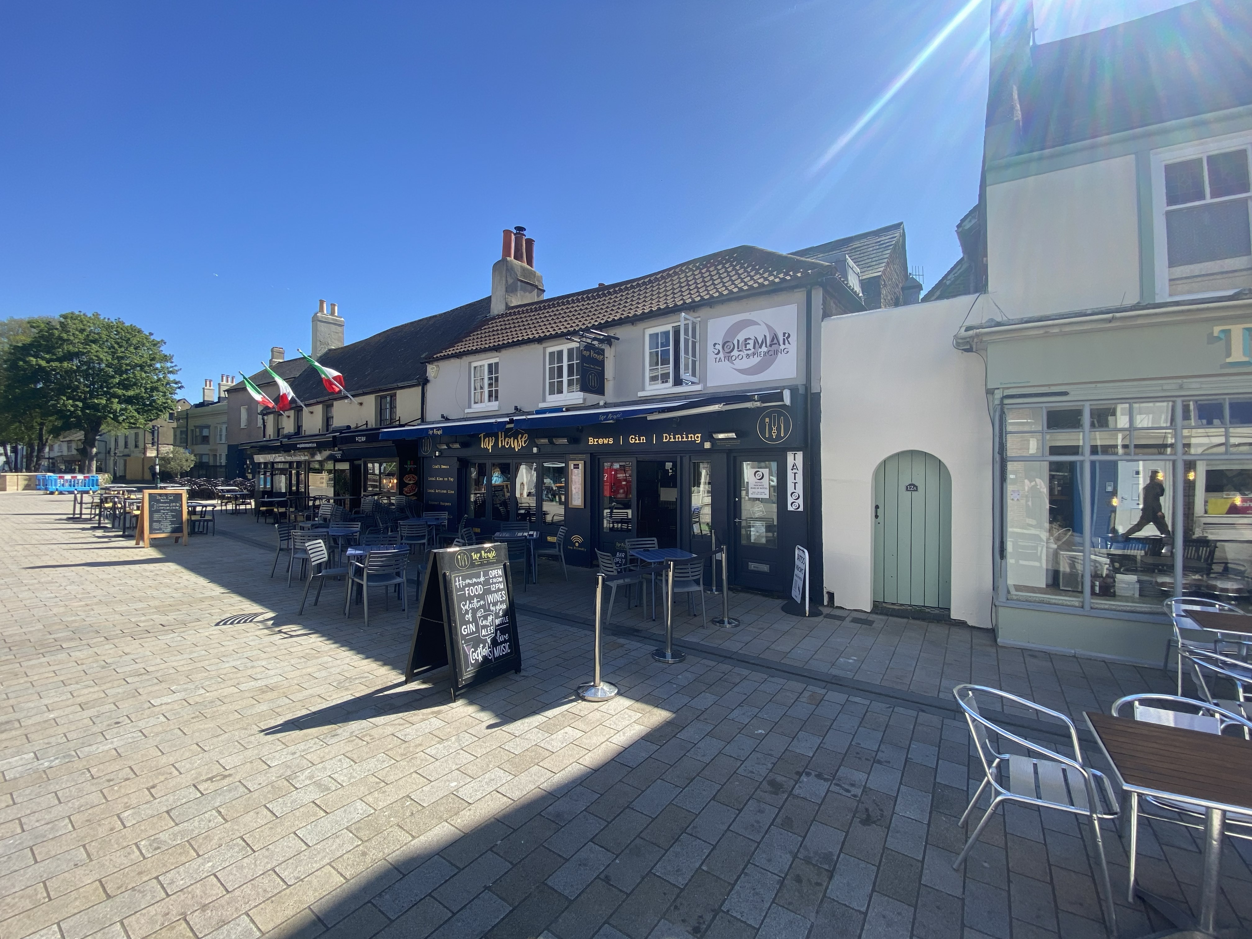Outdoor seating area with tables and chairs in front of Tap House pub and Solemar tattoo and piercing studio under a clear blue sky.