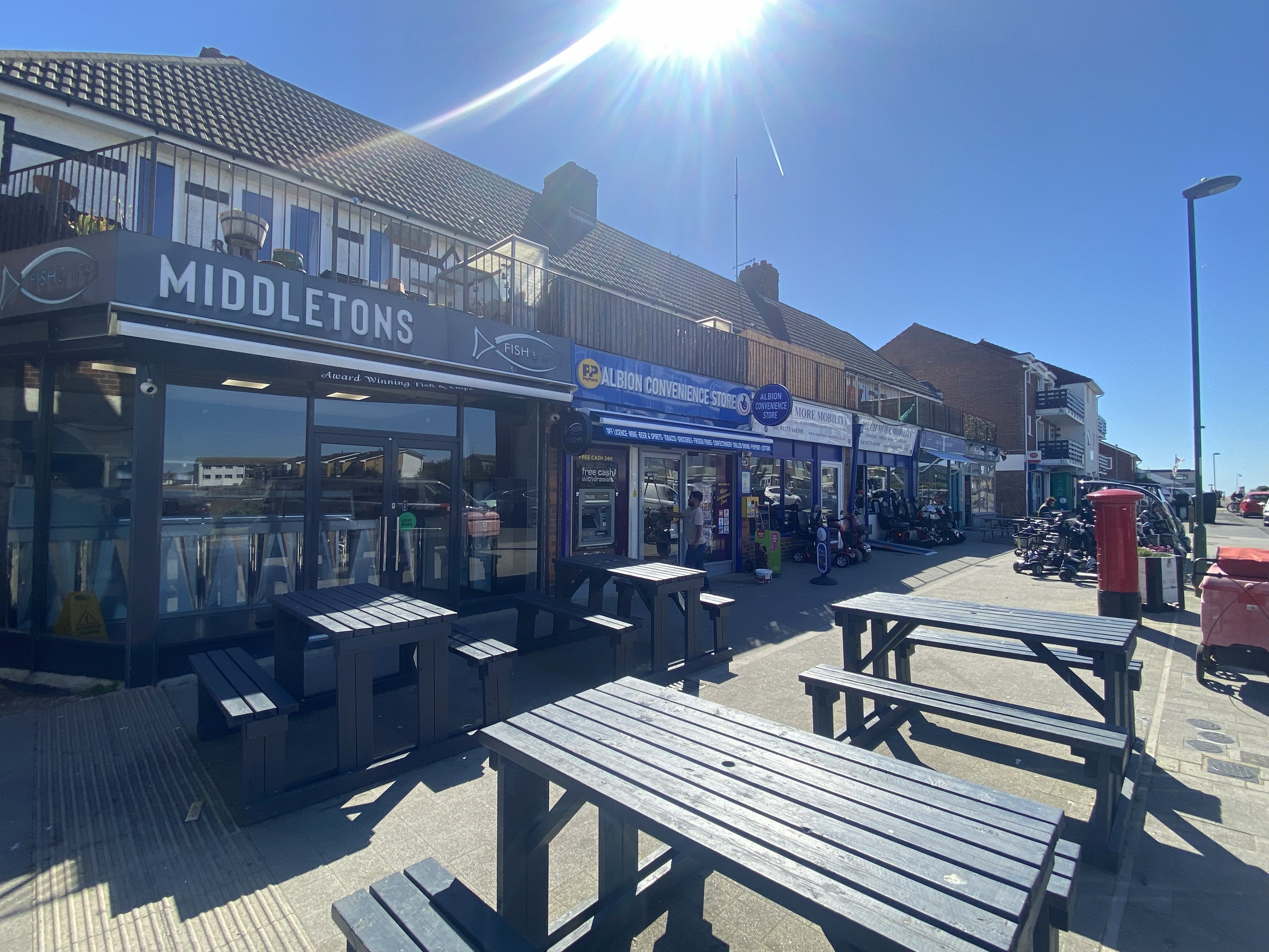 Outdoor view of a street with wooden benches and tables in front of shops including Middletons Fish & Chips and Albion Convenience Store under a bright sunny sky.