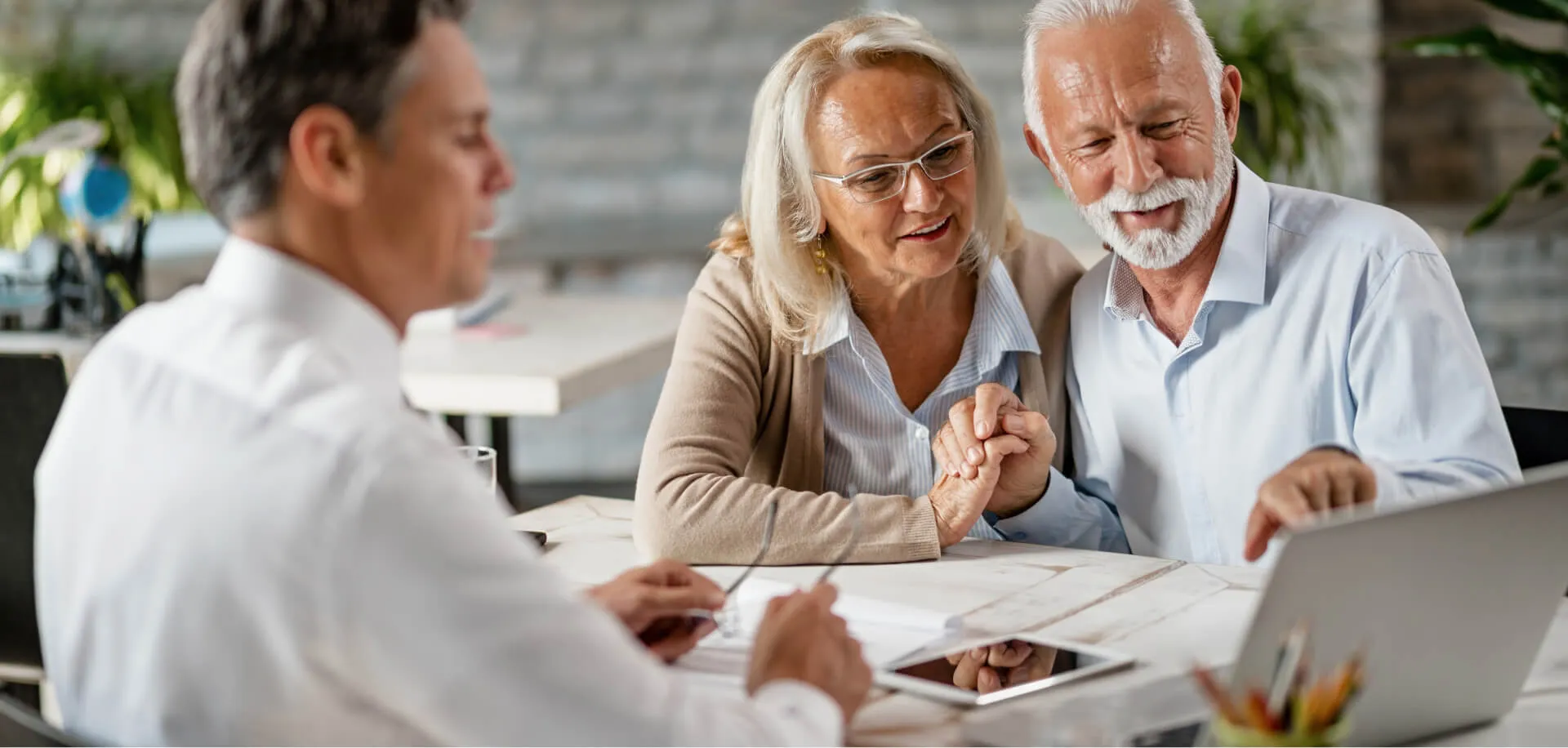Happy senior couple holding hands and using laptop while having a meeting with financial advisor in the office Senior man is pointing at something on laptop