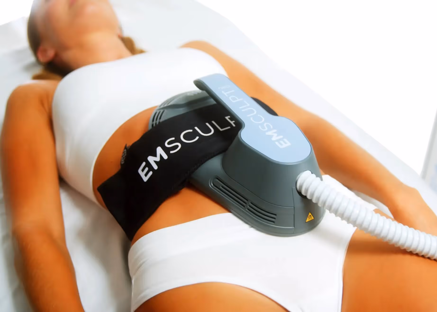 Woman in white sportswear receiving Emsculpt Neo treatment on her abdomen while lying down.